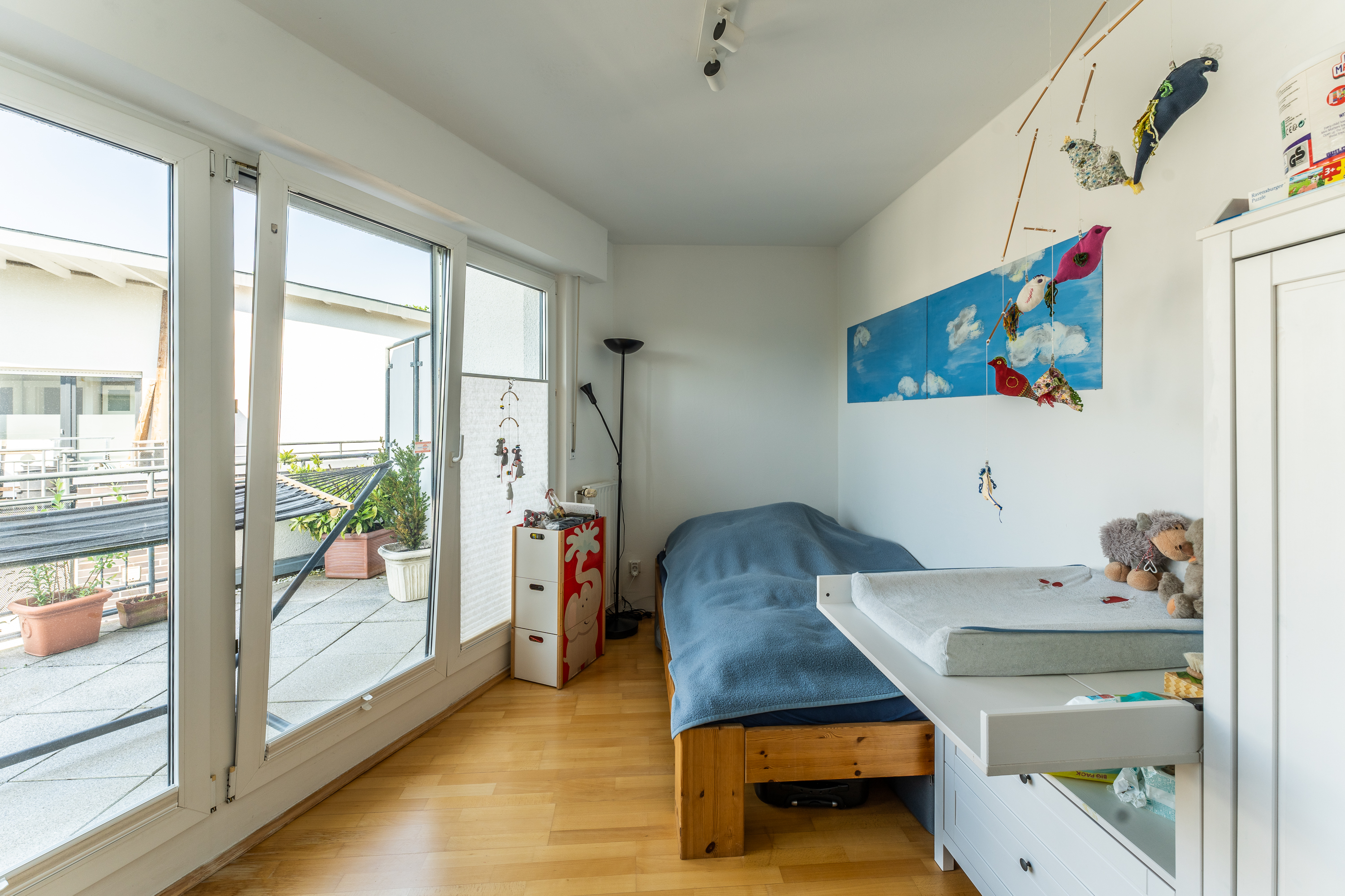 Helles Kinderzimmer mit Bett, Wickelkommode, Spielzeug und Zugang zu einem Balkon mit Pflanzen.