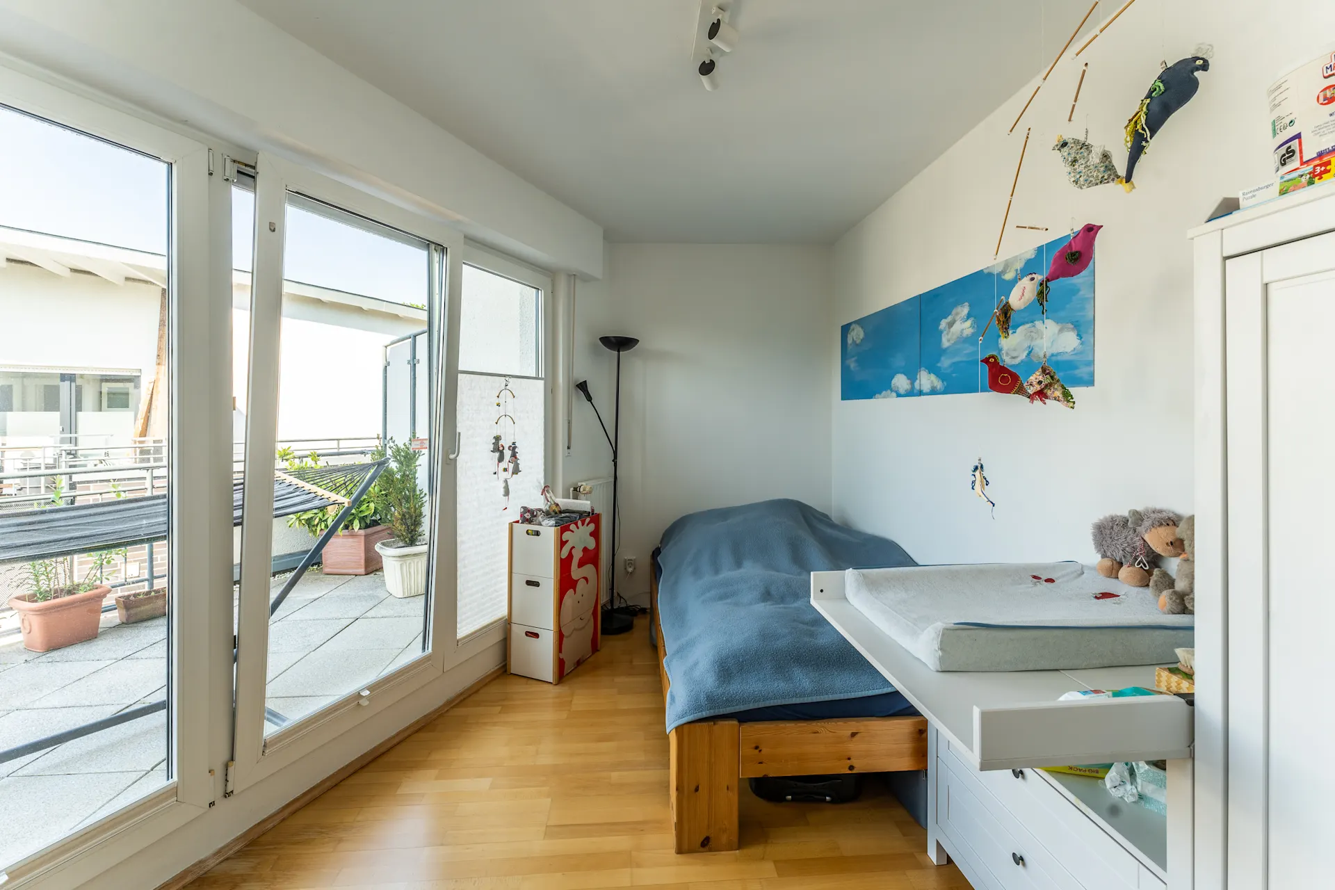 Helles Kinderzimmer mit Bett, Wickelkommode, Spielzeug und Zugang zu einem Balkon mit Pflanzen.