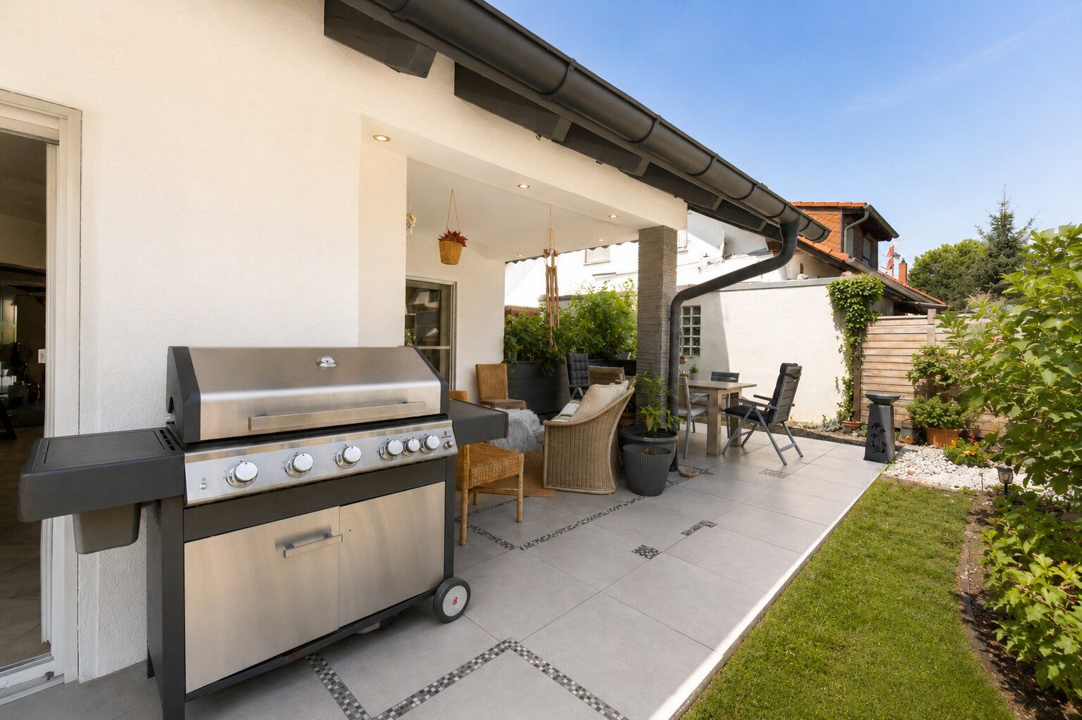 Überdachte Terrasse mit Grill, Sitzmöbeln und Esstisch, umgeben von Grün im Garten.