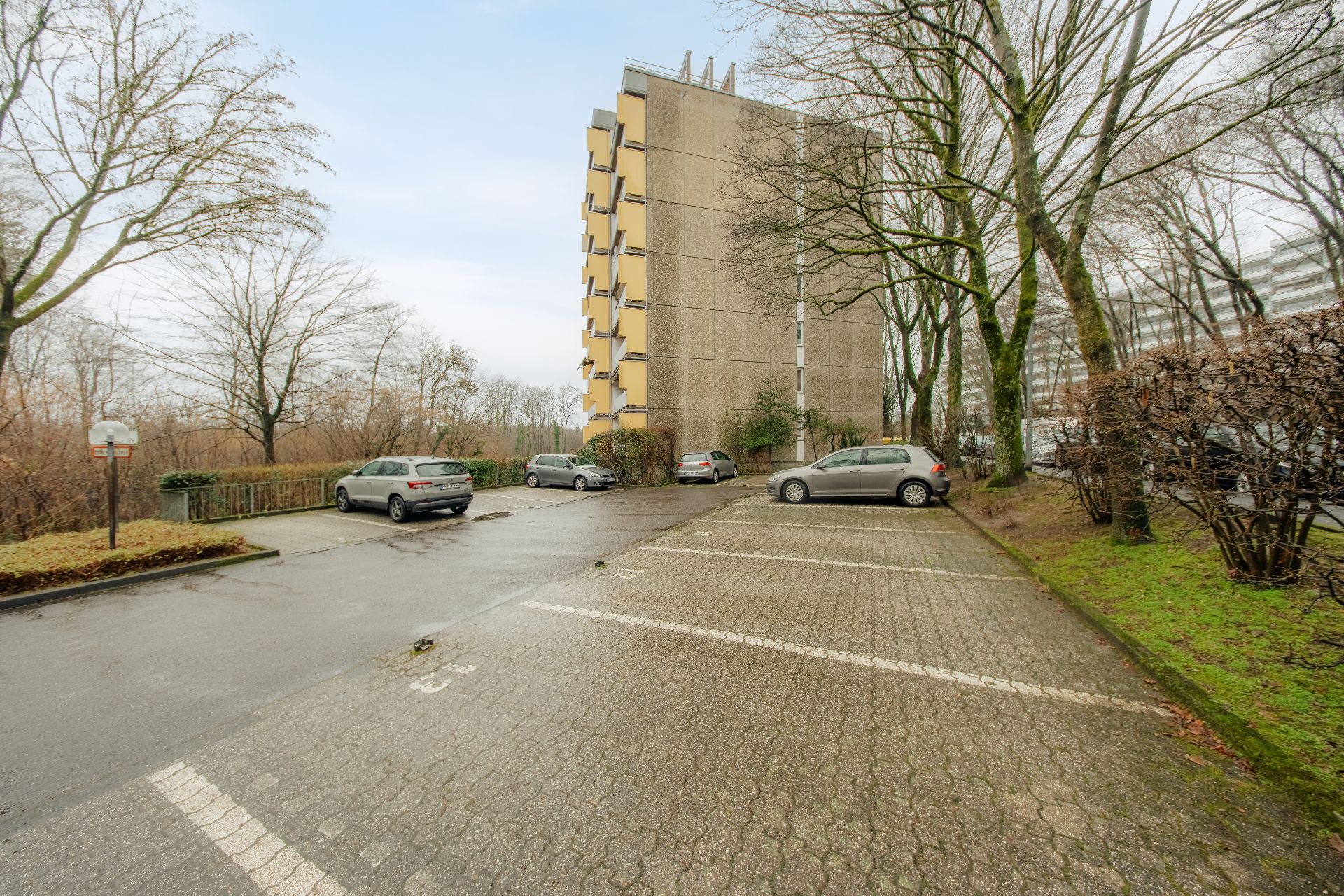 Parkplatz mit mehreren Autos neben einem mehrstöckigen Wohngebäude und Bäumen.