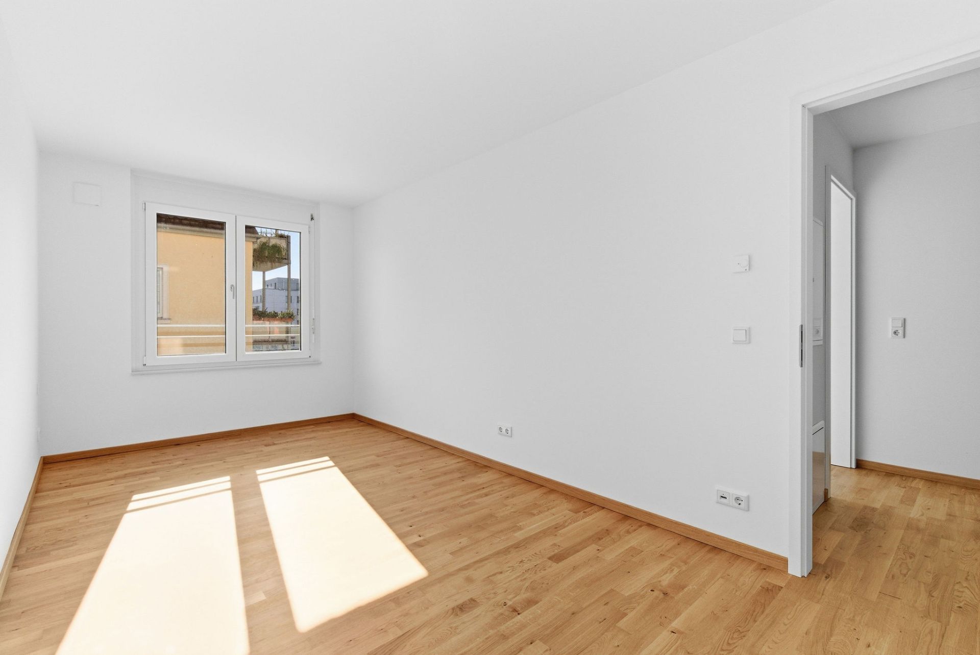 Heller leerer Raum mit Holzfußboden, weißen Wänden und einem Fenster.