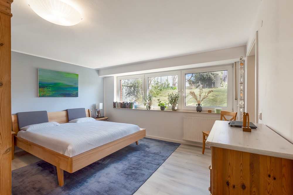 Helles Schlafzimmer mit großem Fenster, Holzbett, Teppich und Kommode.