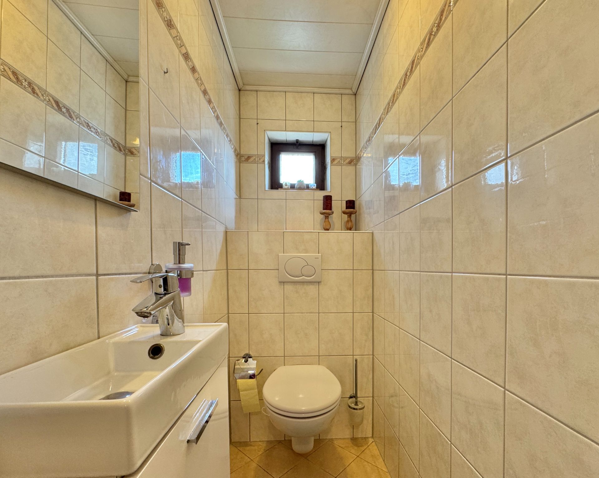 Kleines, gefliestes WC mit Wandtoilette, Waschbecken, Spiegel und kleinem Fenster.
