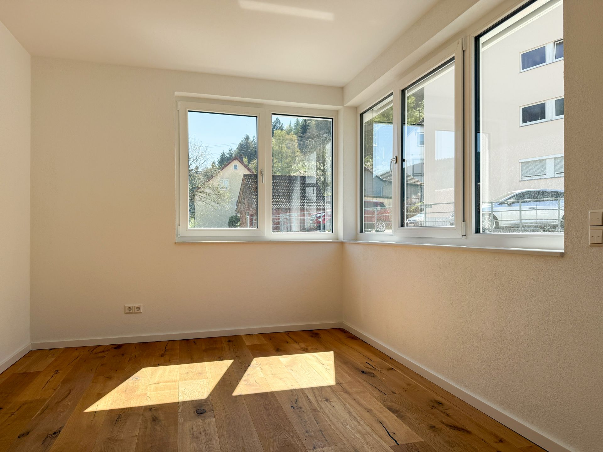 Helles Zimmer mit Holzboden und großen Fenstern an zwei Wänden.