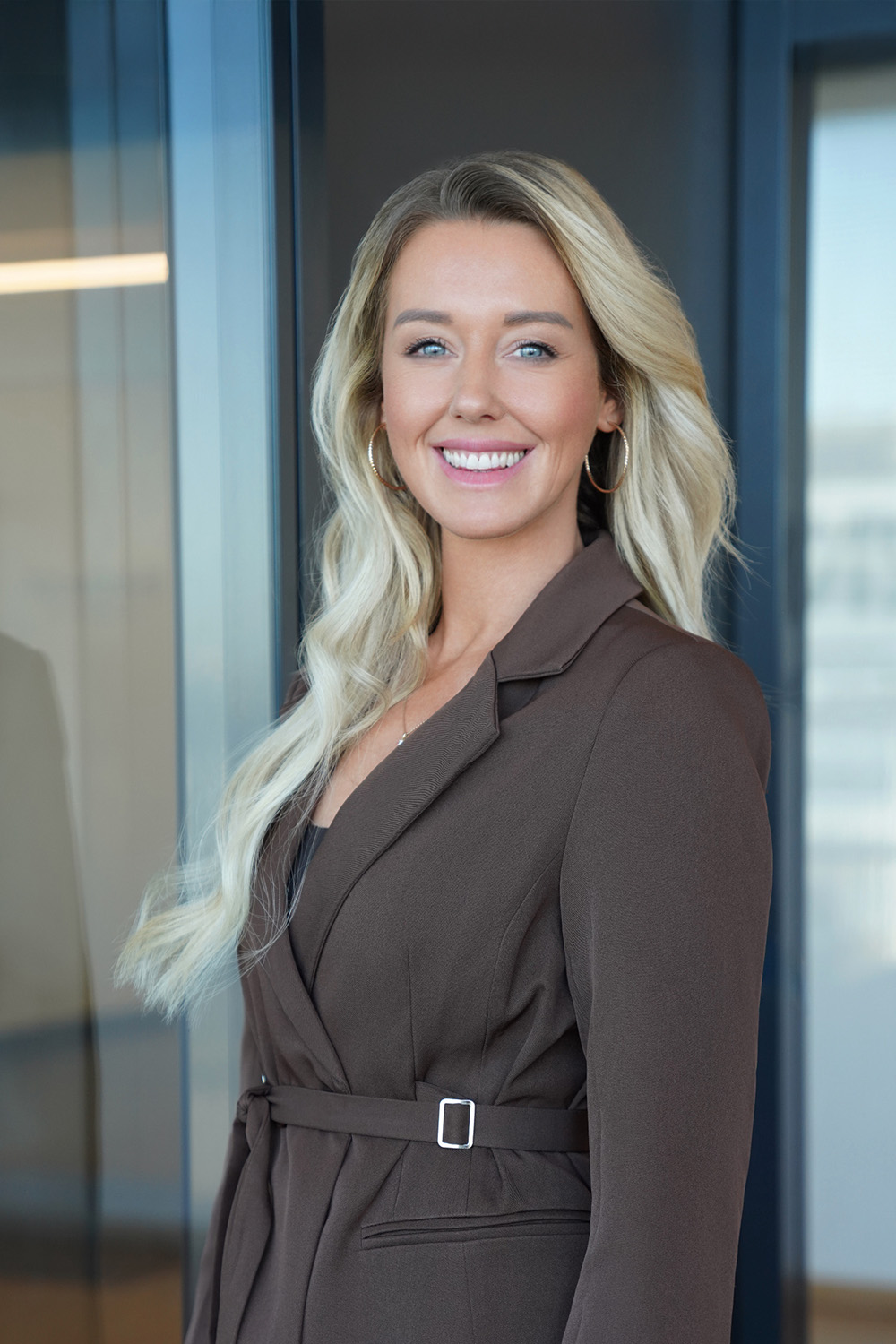 Frau mit langen blonden Haaren und braunem Blazer steht und lächelt in einem modernen Innenraum.