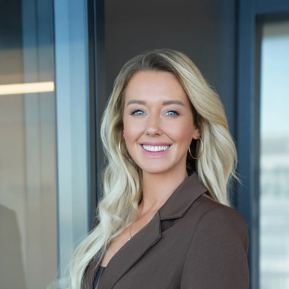 Frau mit langen blonden Haaren und braunem Blazer steht und lächelt in einem modernen Innenraum.