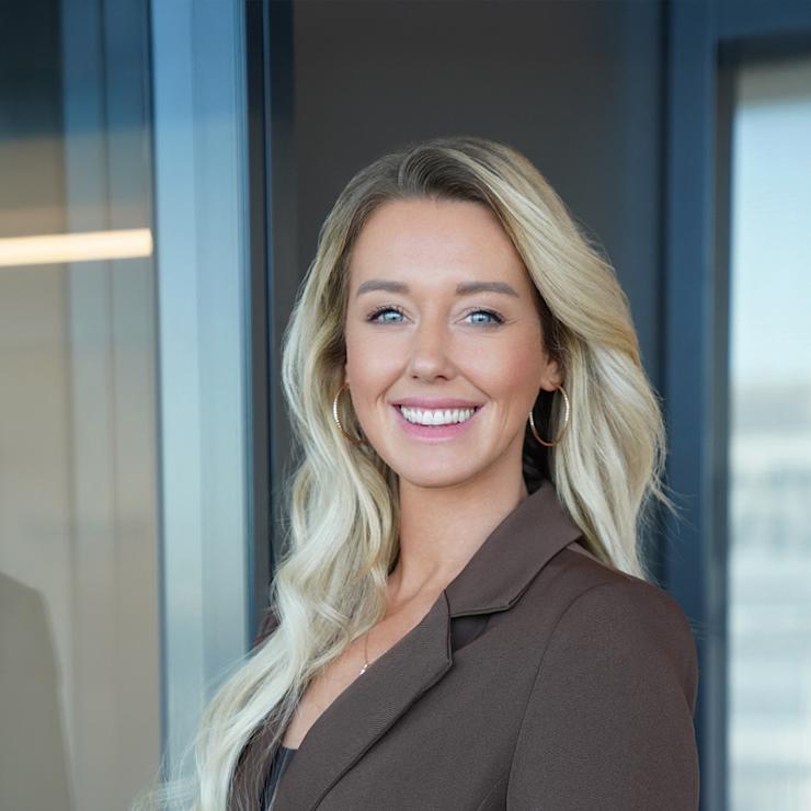 Frau mit langen blonden Haaren und braunem Blazer steht und lächelt in einem modernen Innenraum.