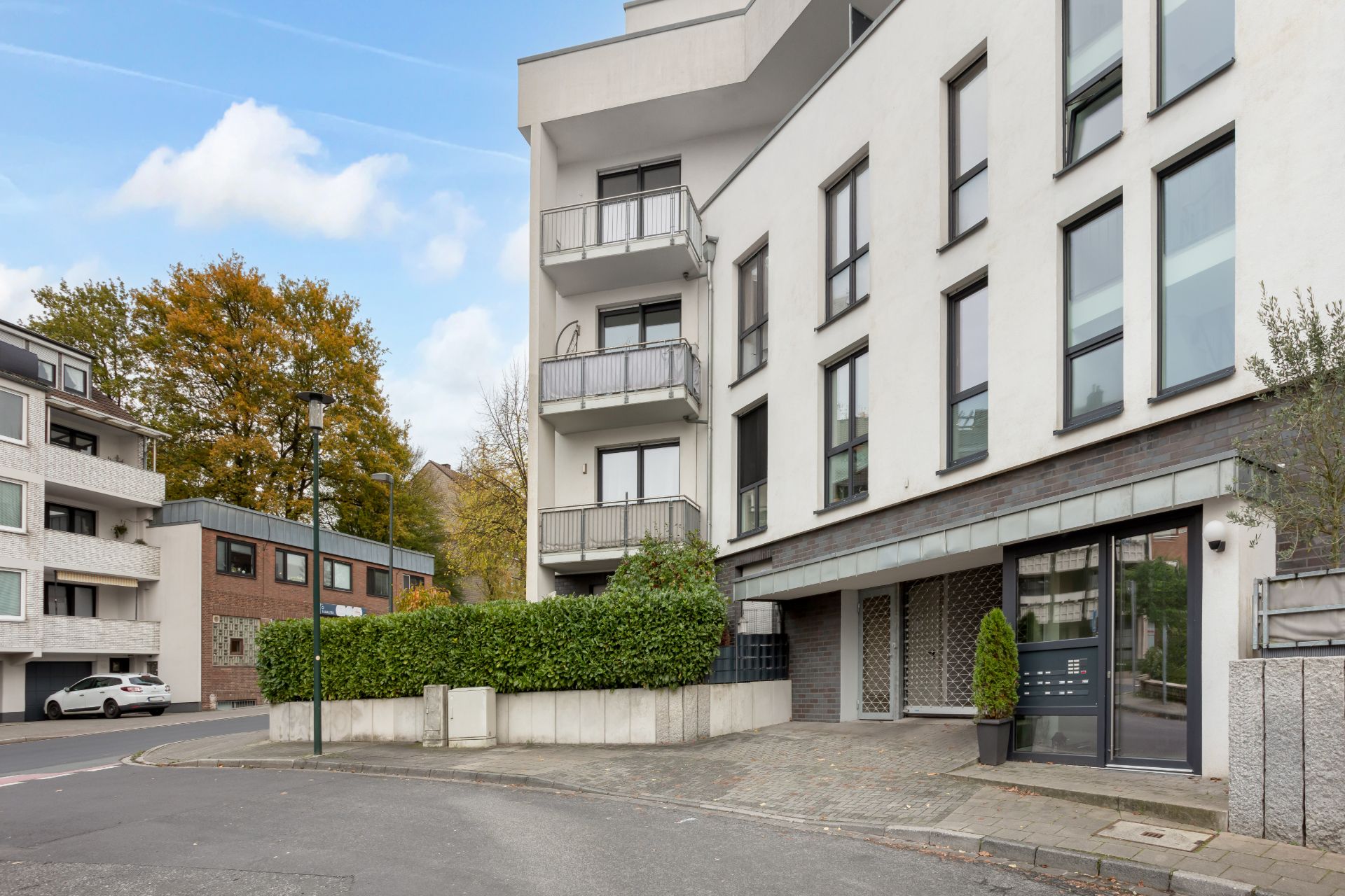 Moderne Wohnhausfassade mit Balkonen, großen Fenstern und einem bepflanzten Eingangsbereich an einer Straßenecke.