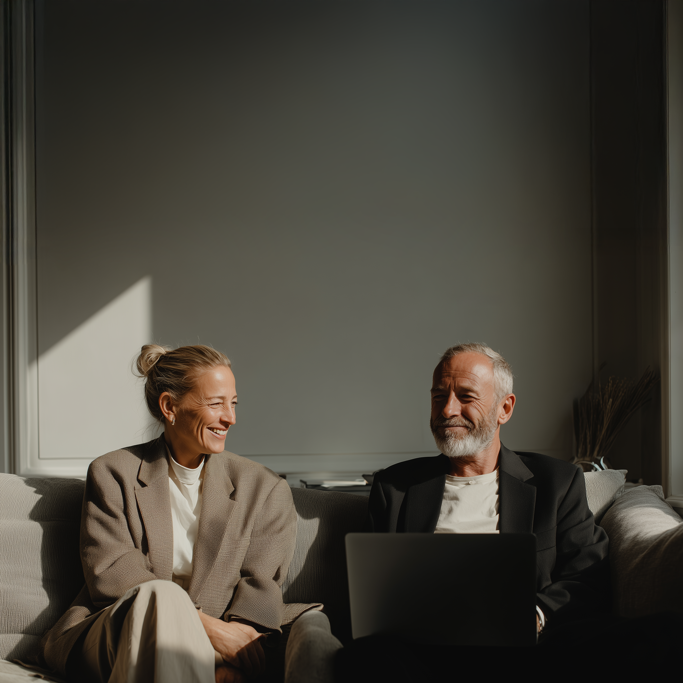 Zwei Menschen sitzen lachend auf einem Sofa mit einem Laptop in einem hellen Wohnzimmer.