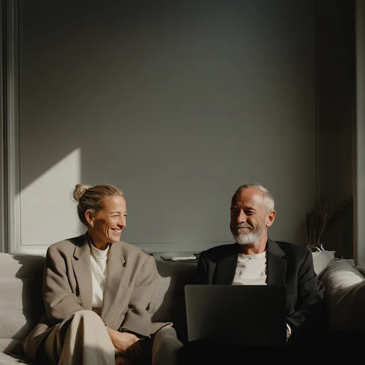 Zwei Menschen sitzen lachend auf einem Sofa mit einem Laptop in einem hellen Wohnzimmer.