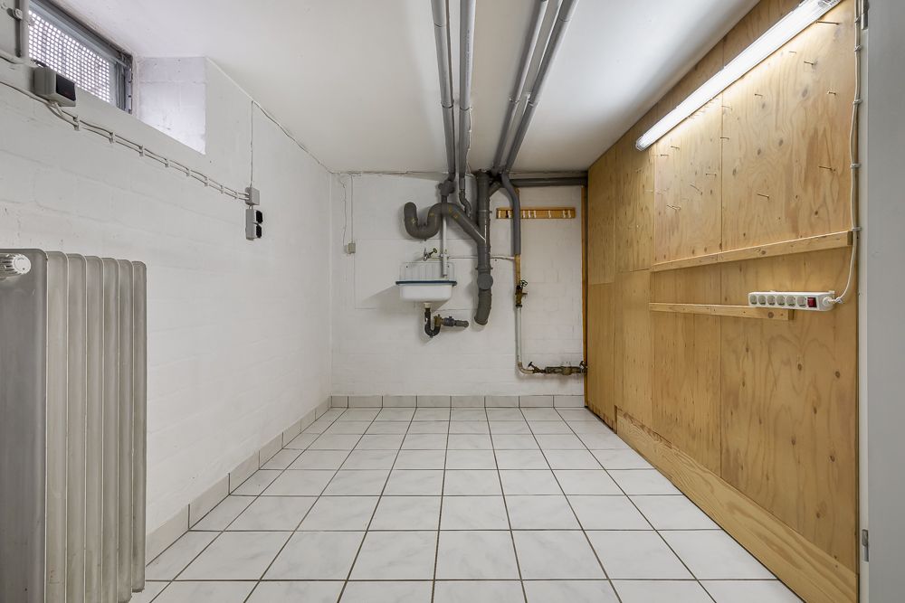 Kellerraum mit weiß gefliesten Boden, Waschbecken, Heizkörper und sichtbaren Rohren.