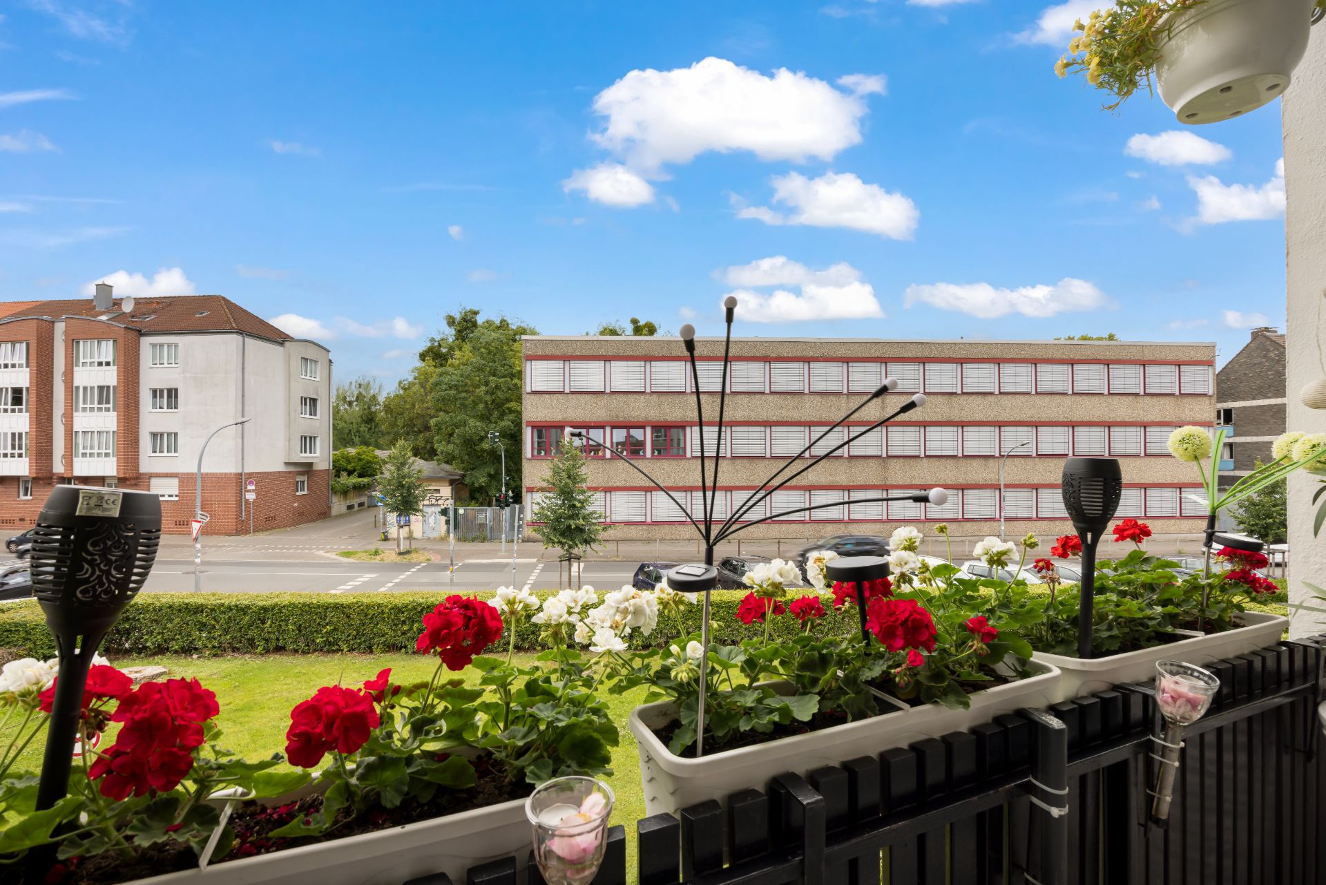 Blick von einem Balkon mit Blumen auf eine Straße und gegenüberliegende Gebäude bei blauem Himmel.
