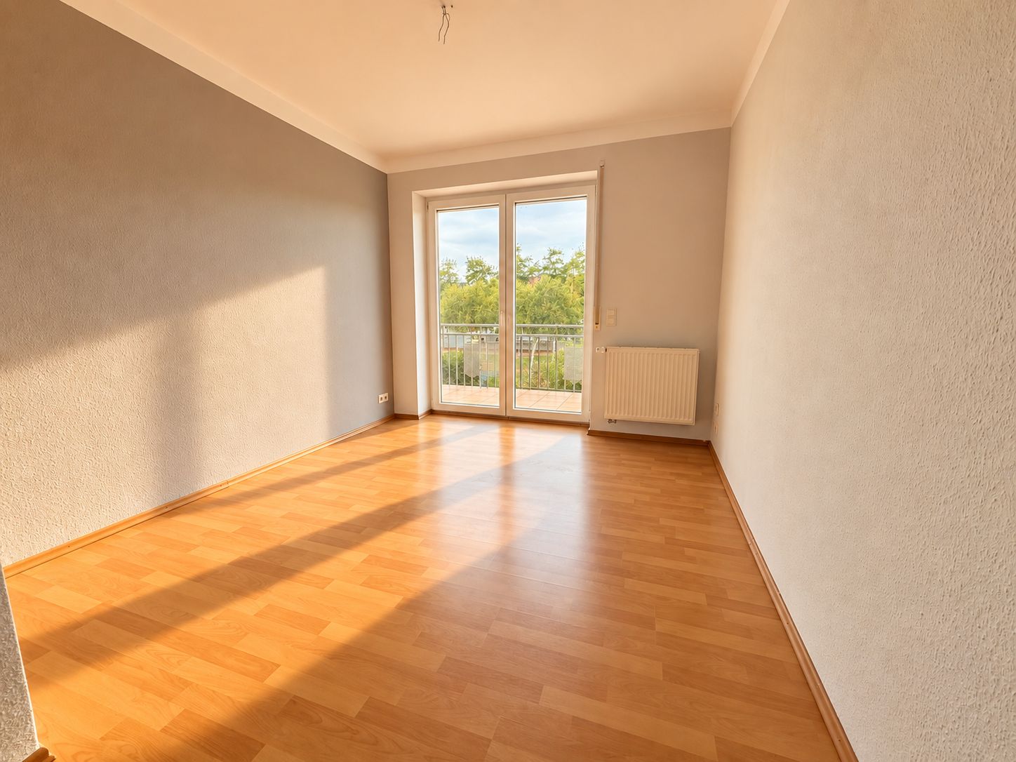 Heller leerer Raum mit Parkettboden, grauen Wänden und großer Glastür zum Balkon.