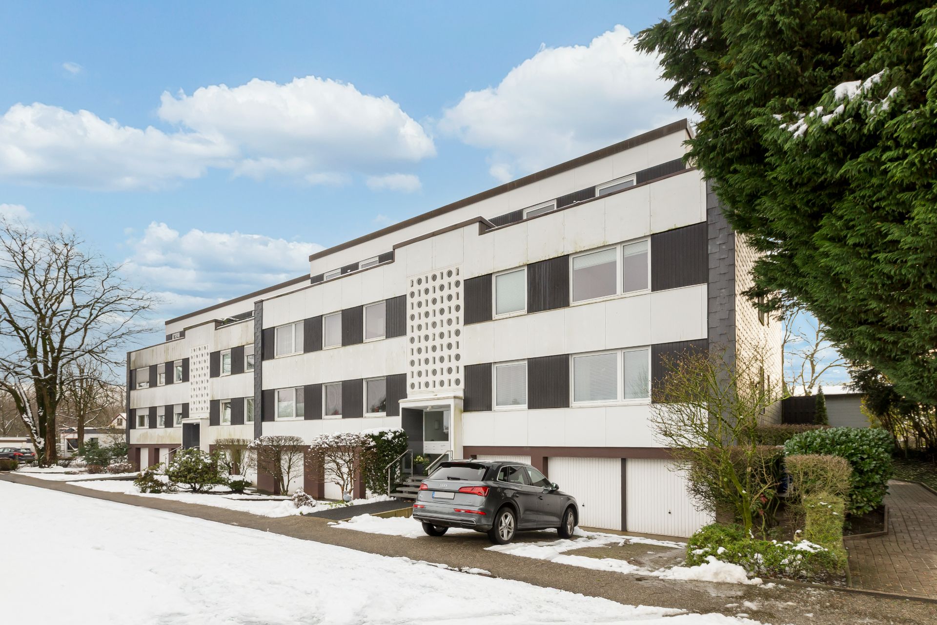 Mehrfamilienhaus mit mehreren Wohnungen, im Winter mit Schnee und parkendem Auto vor dem Eingang.
