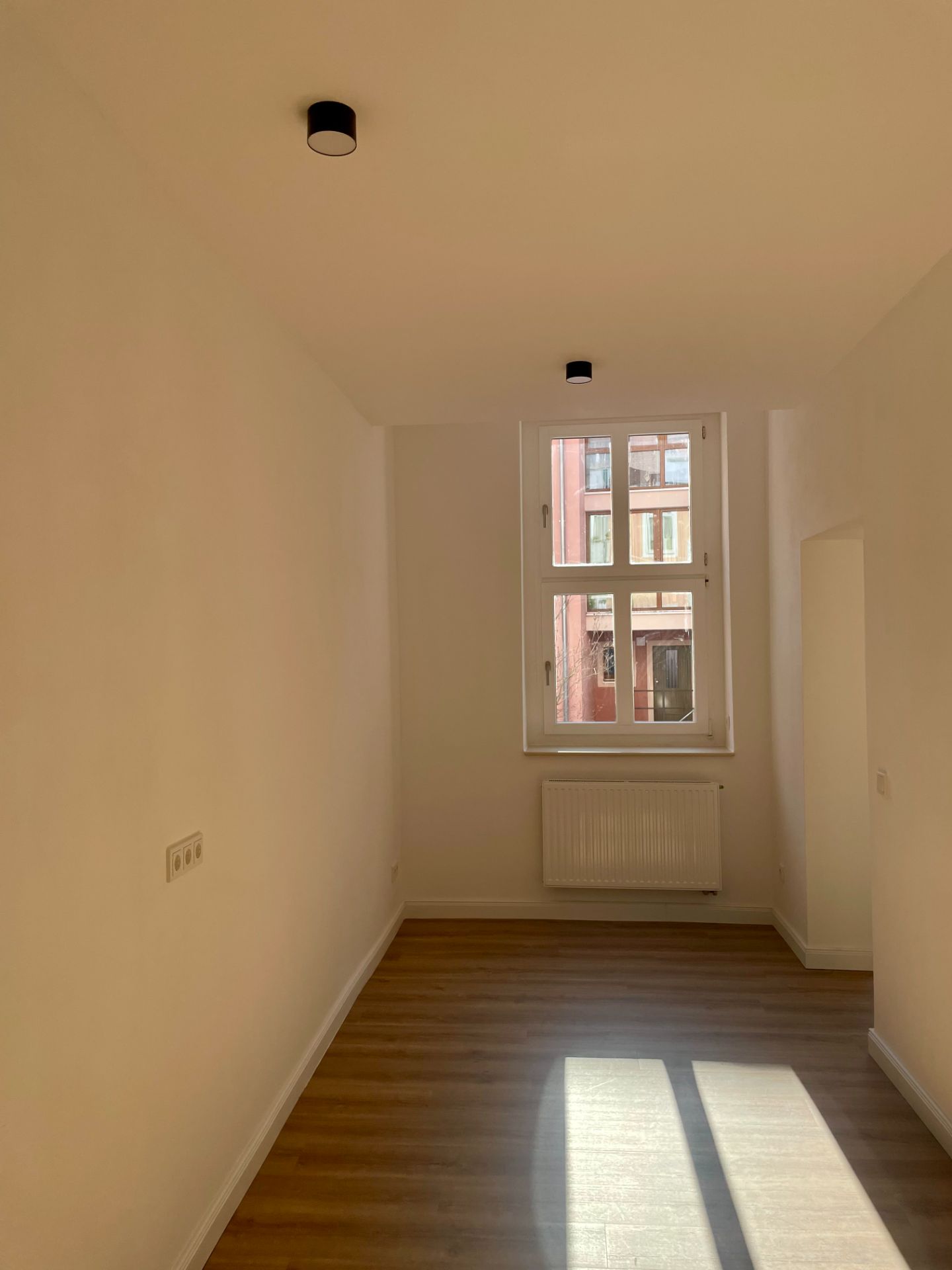 Helles, leeres Zimmer mit Holzfußboden, Fenster und Heizkörper unter dem Fenster.