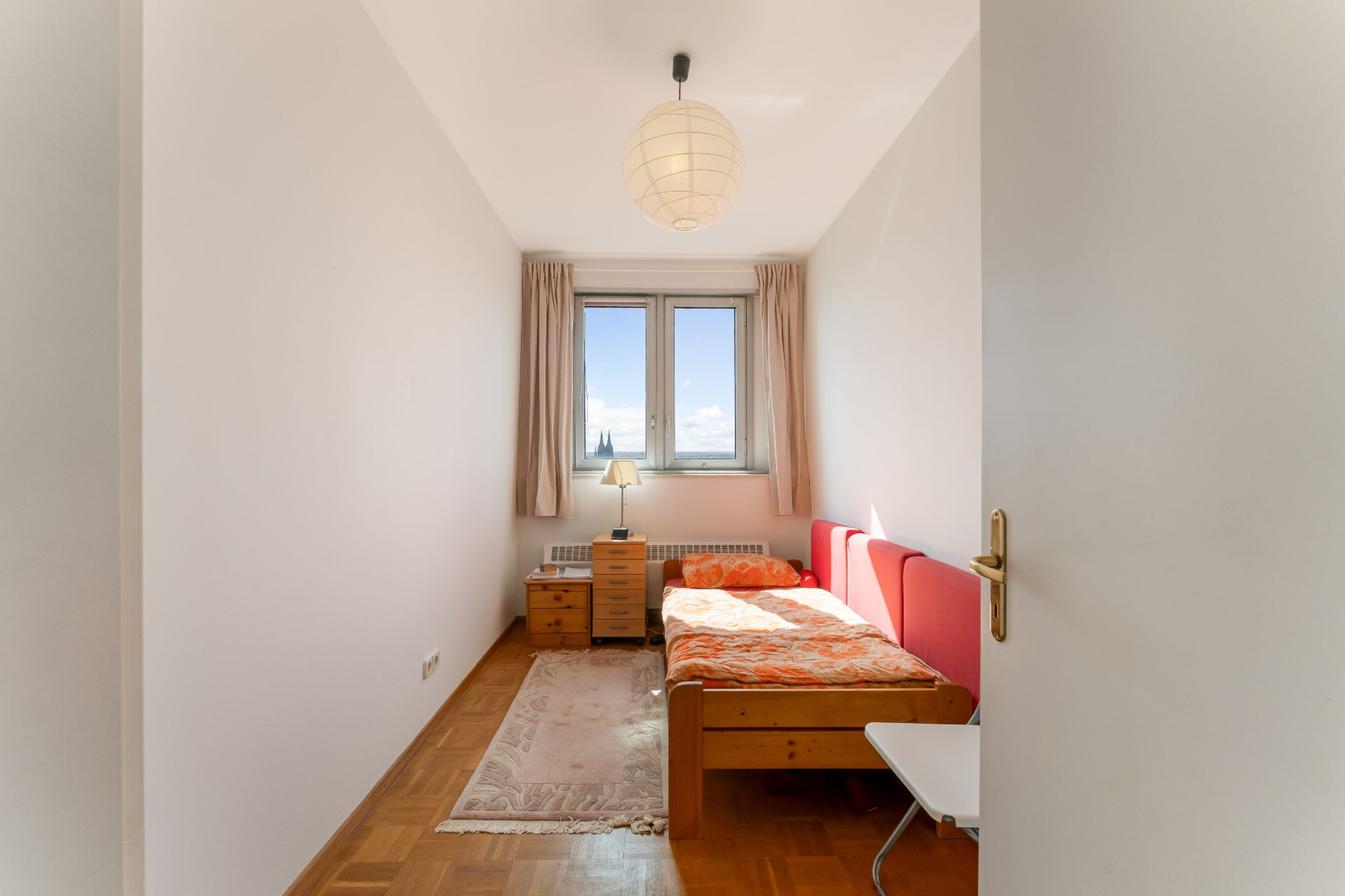 Kleines Schlafzimmer mit Einzelbett, Nachttisch, Lampe und Fenster mit Vorhängen.