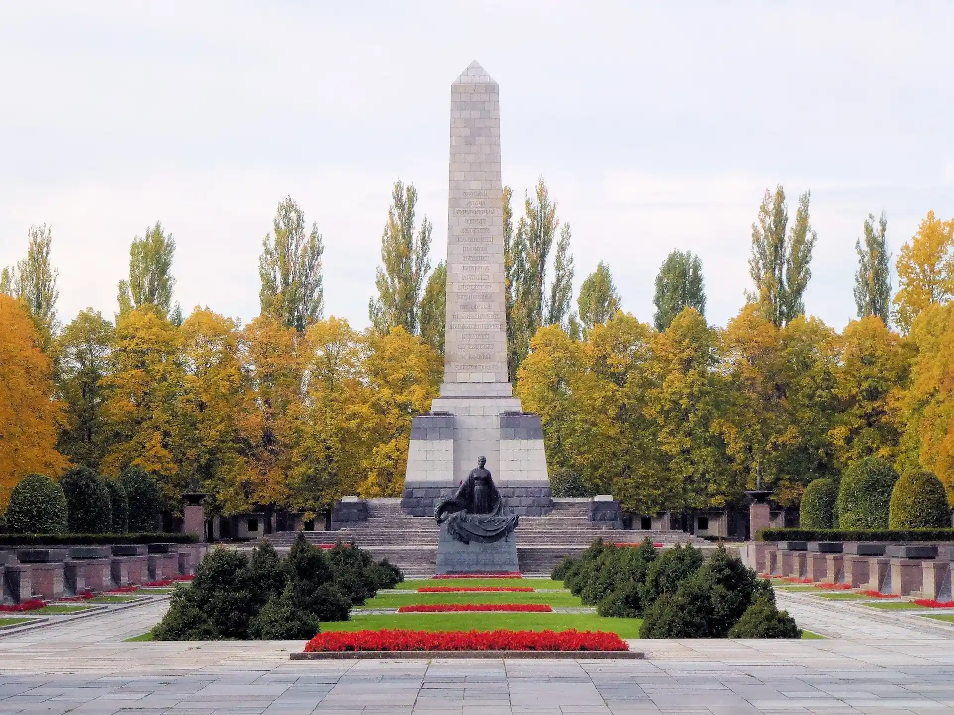 Großes steinernes Denkmal mit Statue und Blumenbeeten, umgeben von Bäumen im Herbst.
