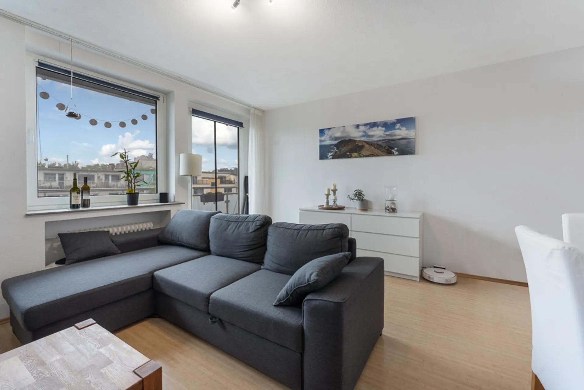 Helles Wohnzimmer mit grauem Ecksofa, Kommode, großem Fenster und Holzfußboden.