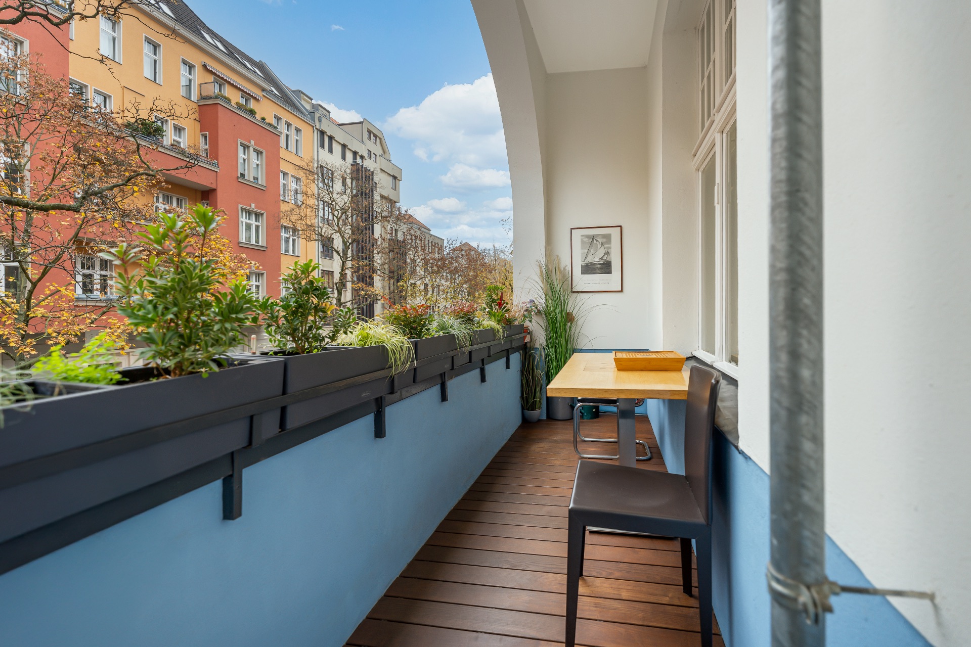 Balkon mit Holzboden, kleinen Pflanzen, Tisch und zwei Stühlen, Blick auf Nachbarhäuser.