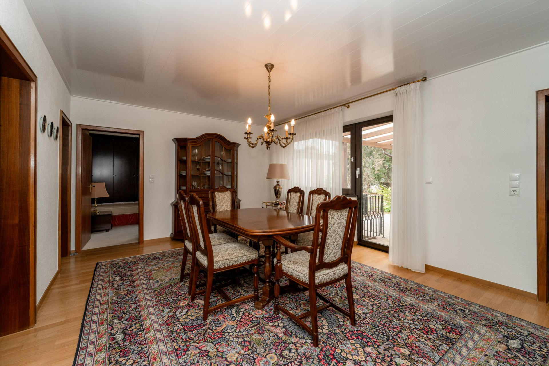 Esszimmer mit Holztisch, sechs Stühlen, Teppich, Kronleuchter und Schrank vor großen Fenstern.