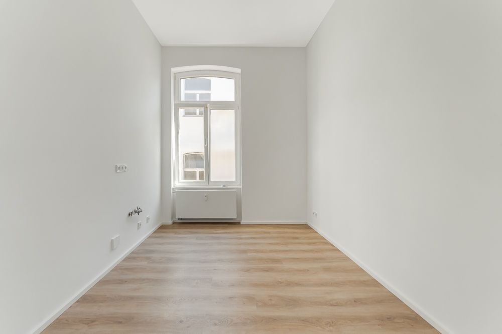 Leerer Raum mit hellen Wänden, Holzfußboden und einem großen Fenster mit Heizung darunter.