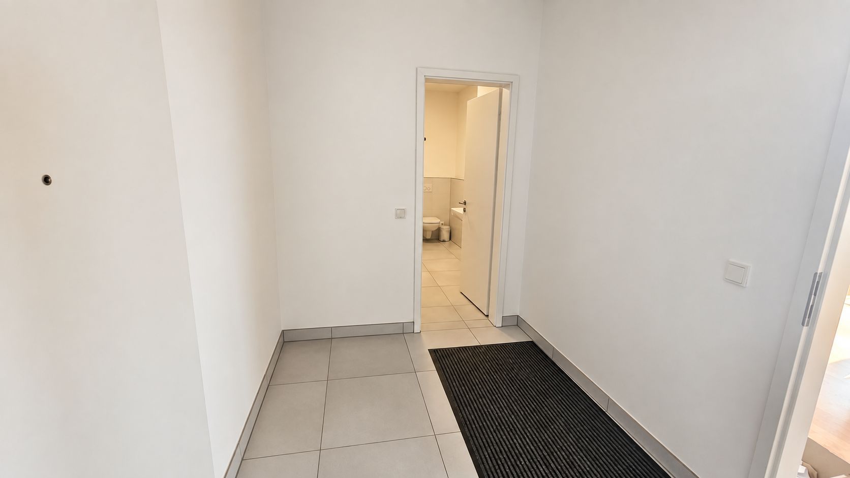 Heller Flur mit Fliesenboden, schwarzer Matte und Blick auf ein offenes Badezimmer mit Toilette.