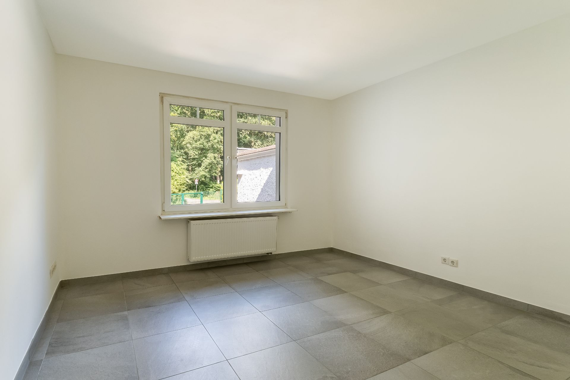 Leerer heller Raum mit großen Fliesen, weißer Wand und Fenster mit Blick ins Grüne.