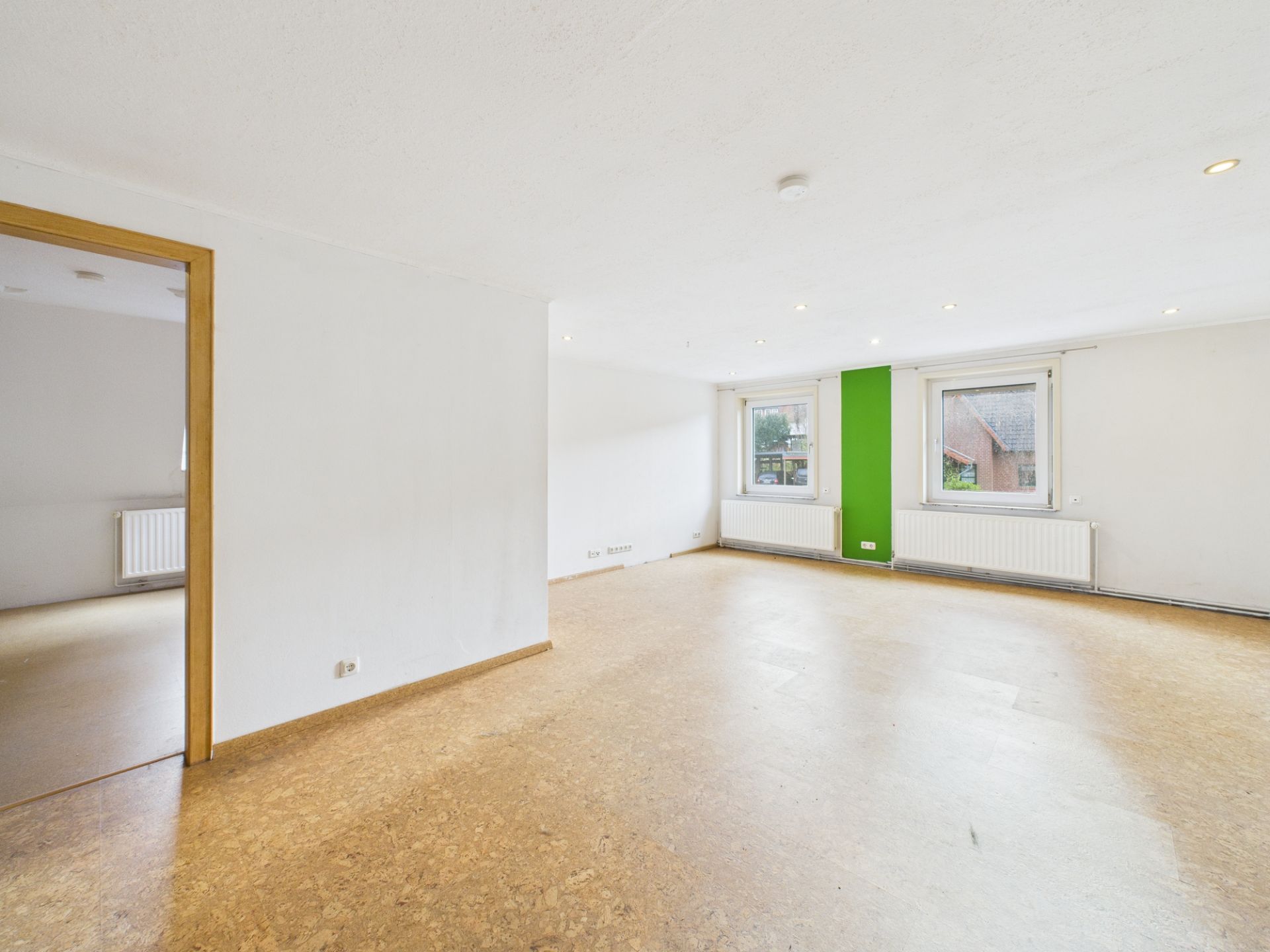Helles, leeres Zimmer mit zwei Fenstern, grüner Wandfläche und offenem Durchgang zu einem weiteren Raum.