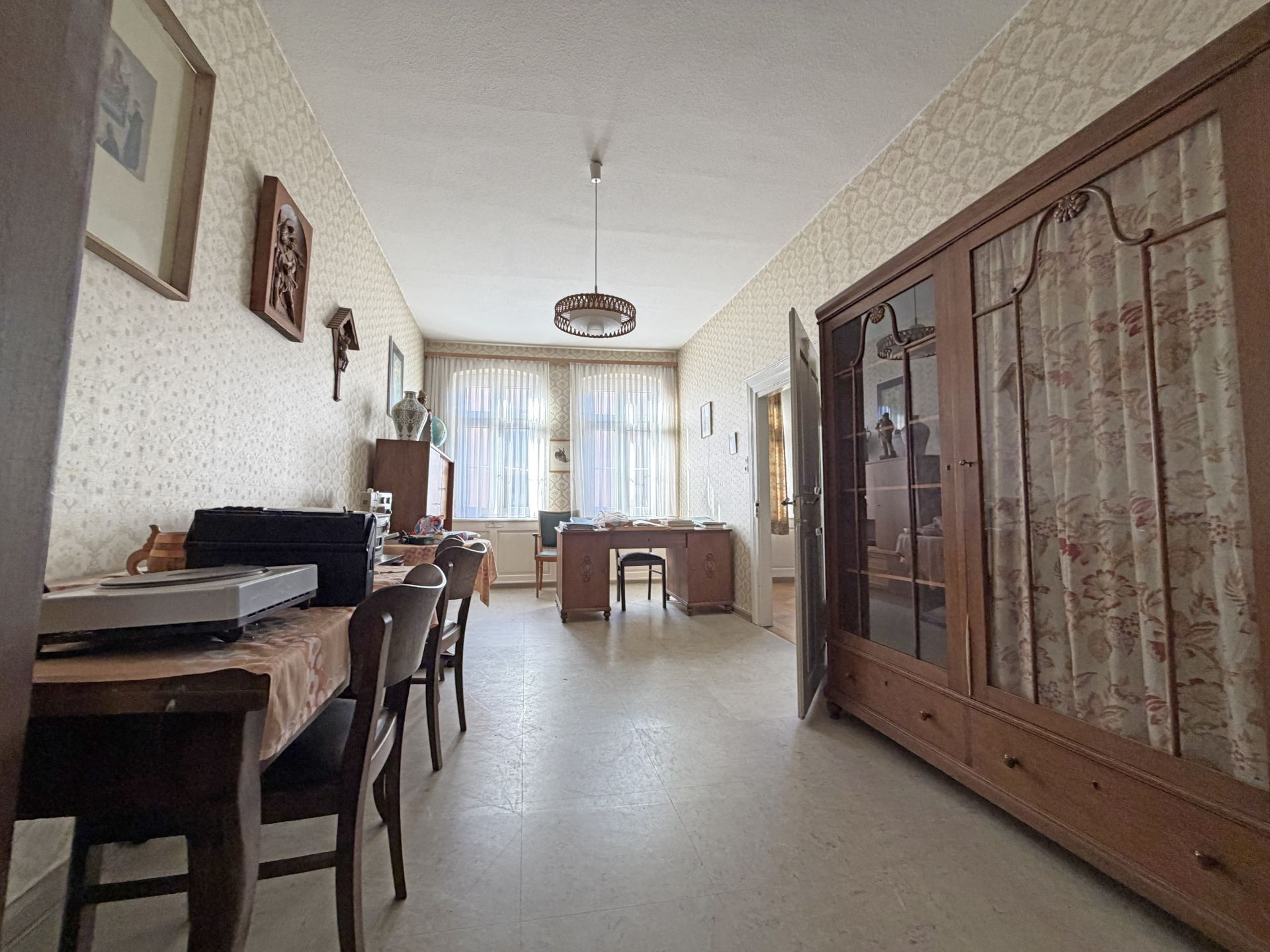 Helles Zimmer mit großem Schrank, Esstisch, Stühlen und Schreibtisch, Fenster im Hintergrund.