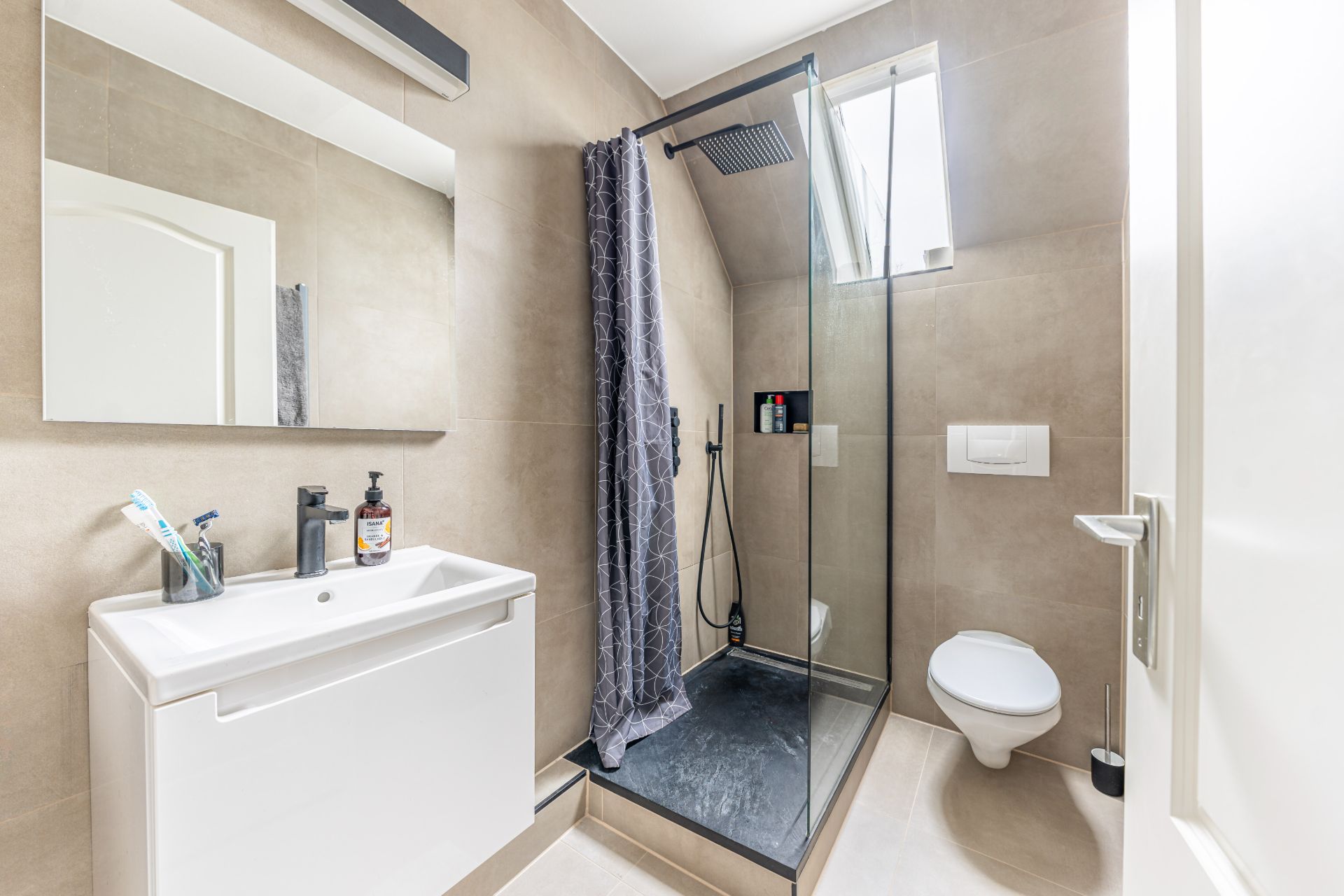 Modernes Badezimmer mit Dusche, Waschbecken und Wandtoilette, beige Fliesen und Fenster.