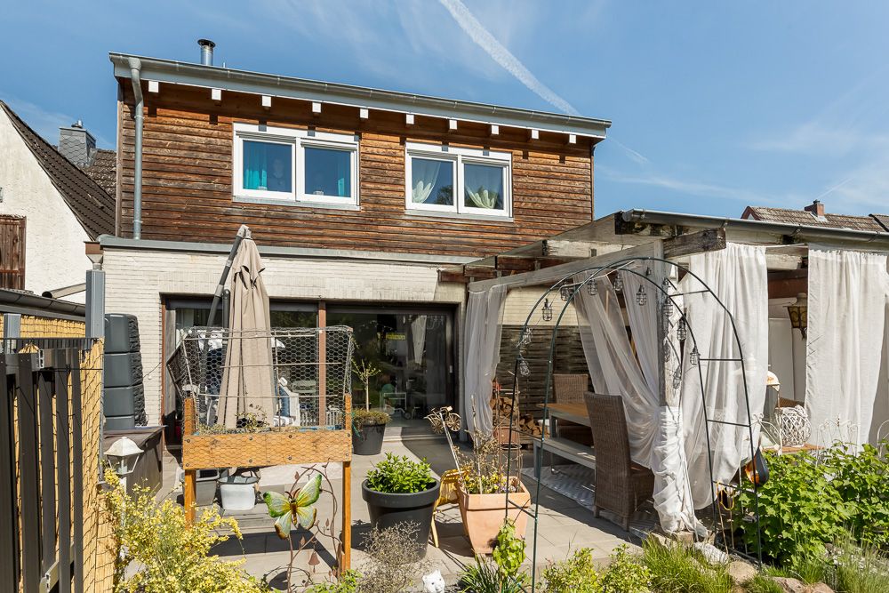 Zweistöckiges Wohnhaus mit Holzfassade, Terrasse, Pflanzen und Gartenmöbeln im Garten.