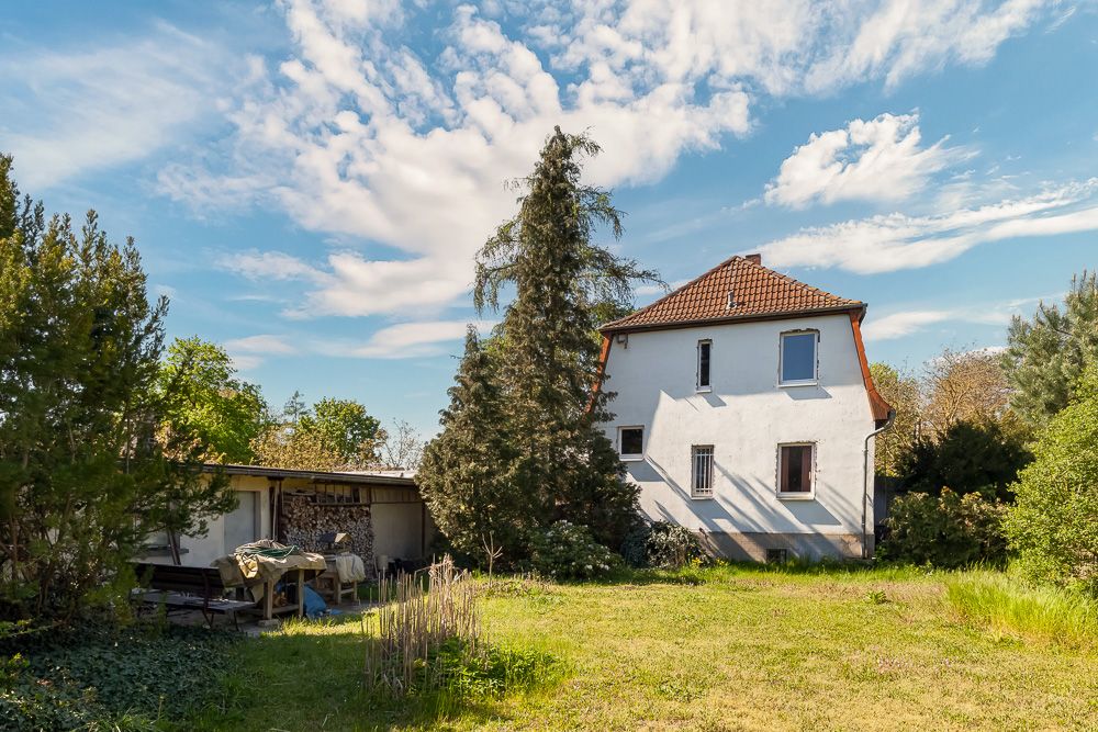 Weißes Wohnhaus mit rotem Dach in einem grünen Garten, Bäume und ein kleiner Schuppen im Hintergrund.