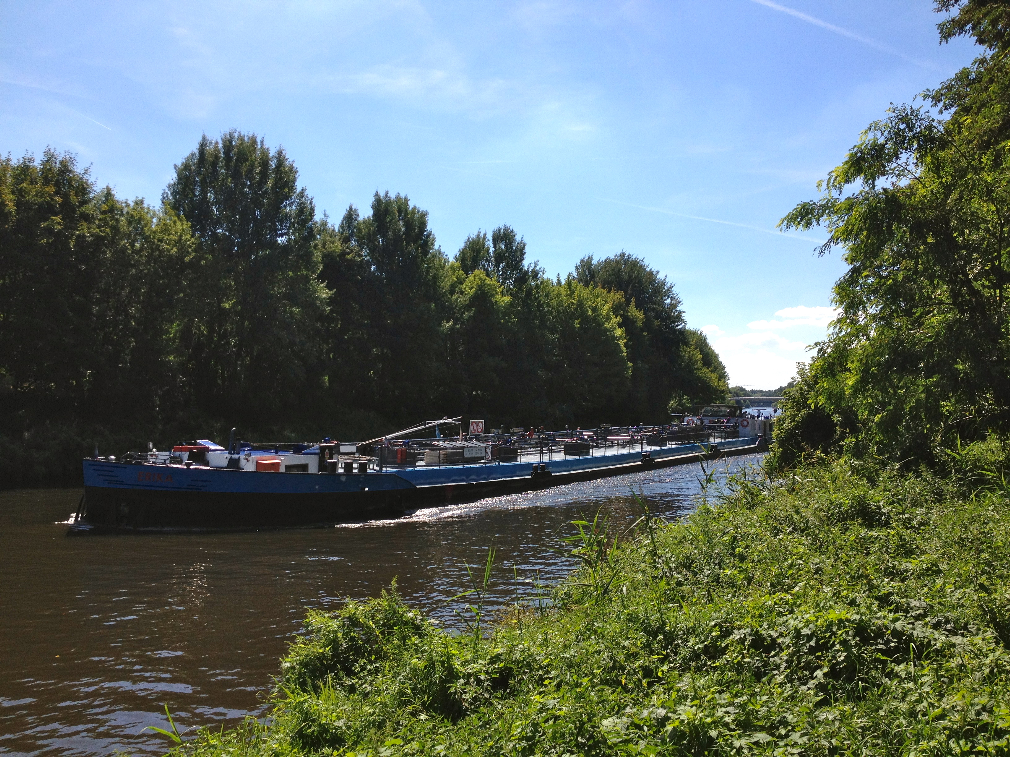 Ein langer Frachter fährt auf einem Fluss, umgeben von grünen Bäumen bei sonnigem Wetter.
