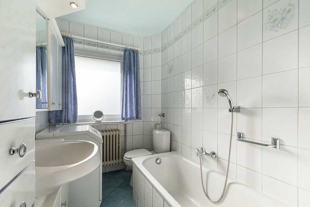 Helles Badezimmer mit Badewanne, Waschbecken, Toilette und Fenster mit blauen Vorhängen.