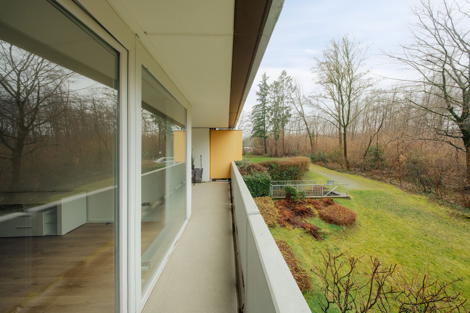 Langer Balkon mit Glasgeländer, Blick auf Garten und Bäume im Hintergrund.
