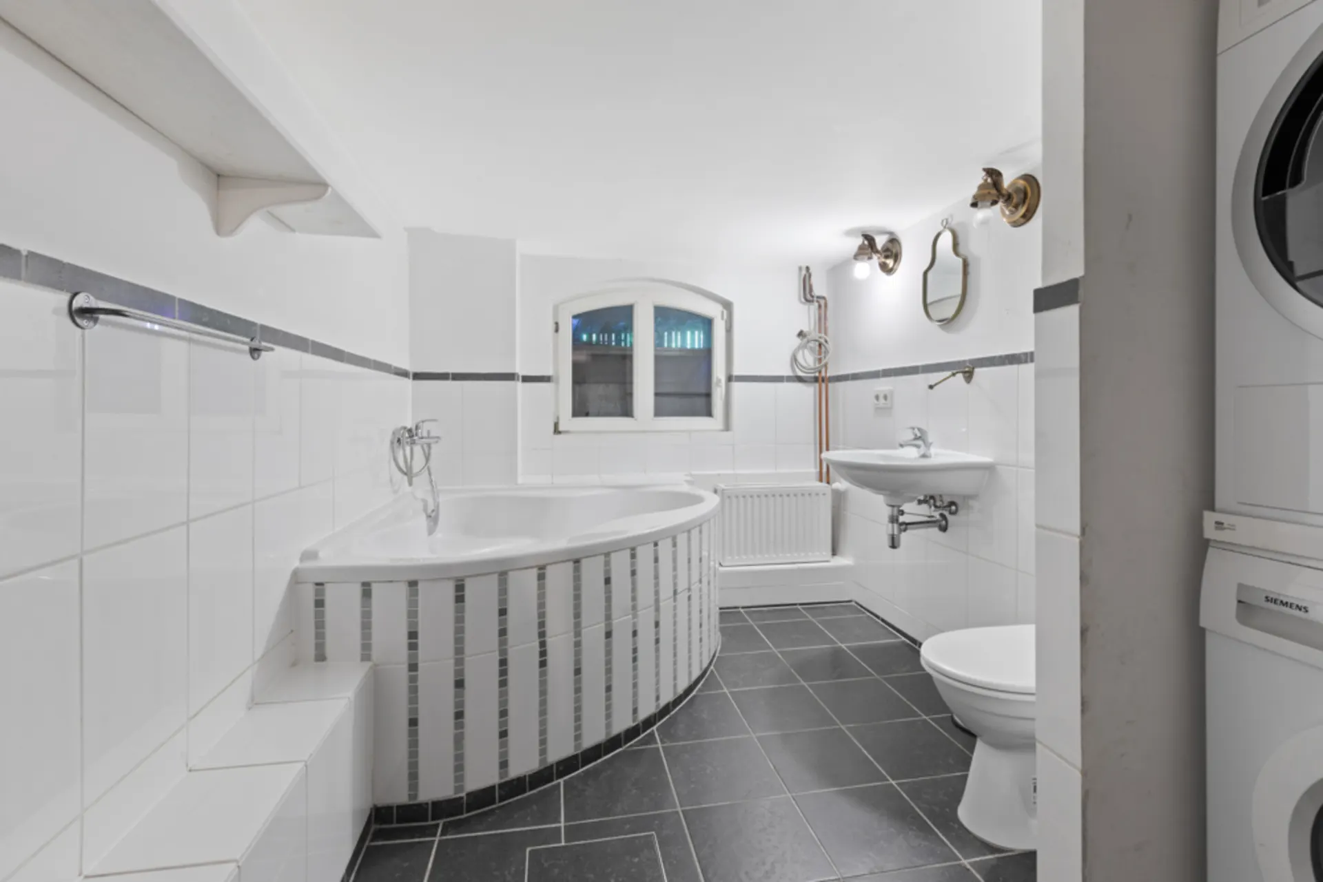 Modernes Badezimmer mit Eckbadewanne, Waschbecken, Toilette und Stapelwaschmaschine.