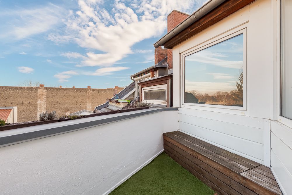Kleine Dachterrasse mit Kunstrasen, Holzbank und Blick auf Hauswände bei blauem Himmel.