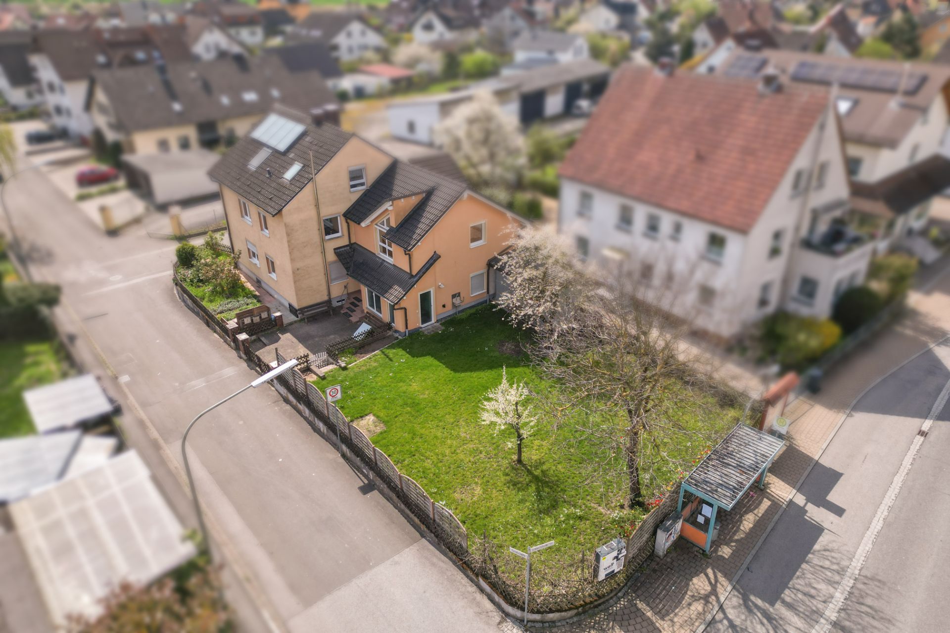 Luftaufnahme eines Wohnhauses mit umzäuntem Garten und zwei Bäumen an einer Straßenecke.