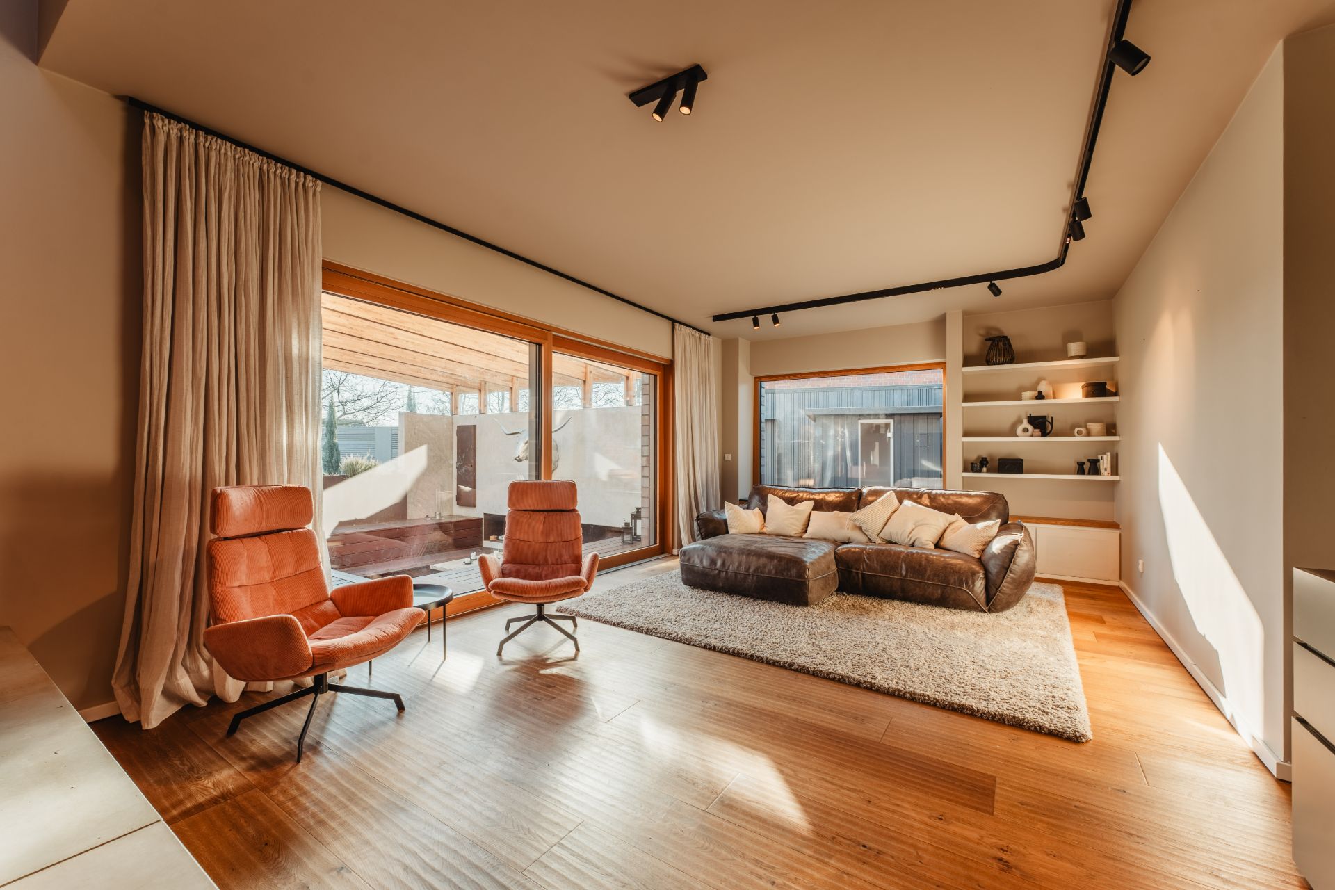 Modernes Wohnzimmer mit braunem Sofa, zwei orange Sesseln und großen Fenstern.
