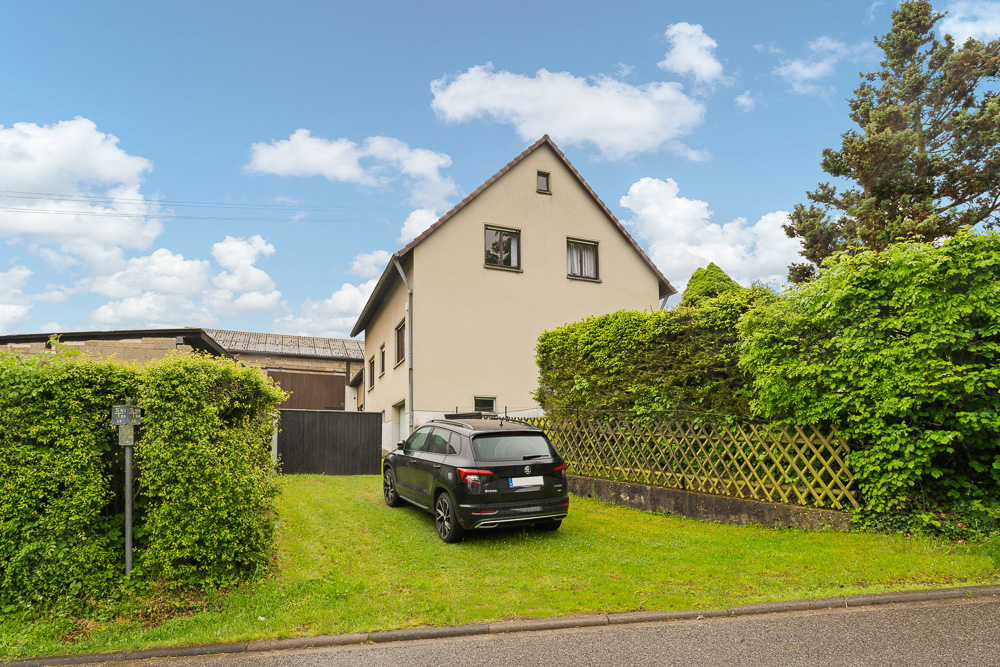 Haus mit Satteldach, schwarzes Auto auf Rasen, umgeben von Hecken und Bäumen.