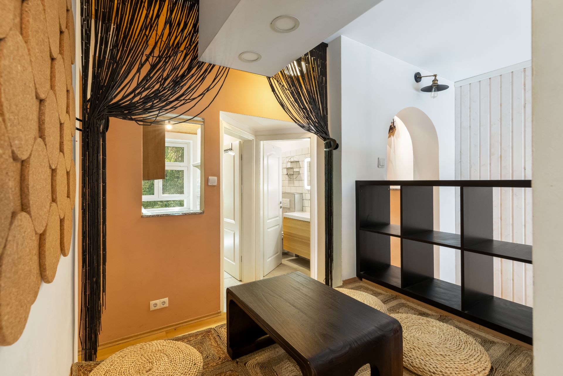Kleiner Wohnraum mit niedrigem Tisch, Regal, runden Teppichen und Blick ins Badezimmer.