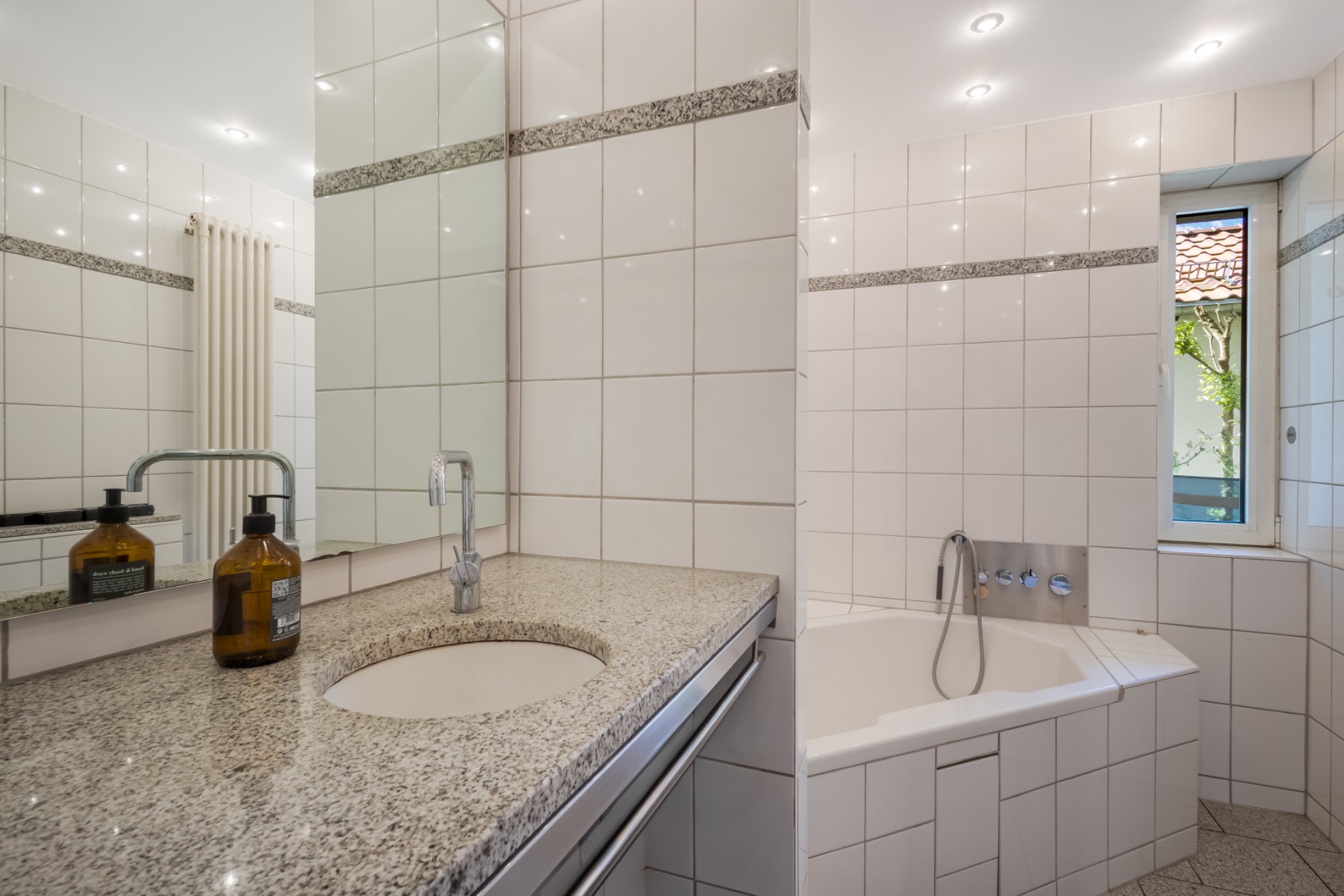 Modernes, hell gefliestes Badezimmer mit Waschbecken, Spiegel, Badewanne und Fenster.