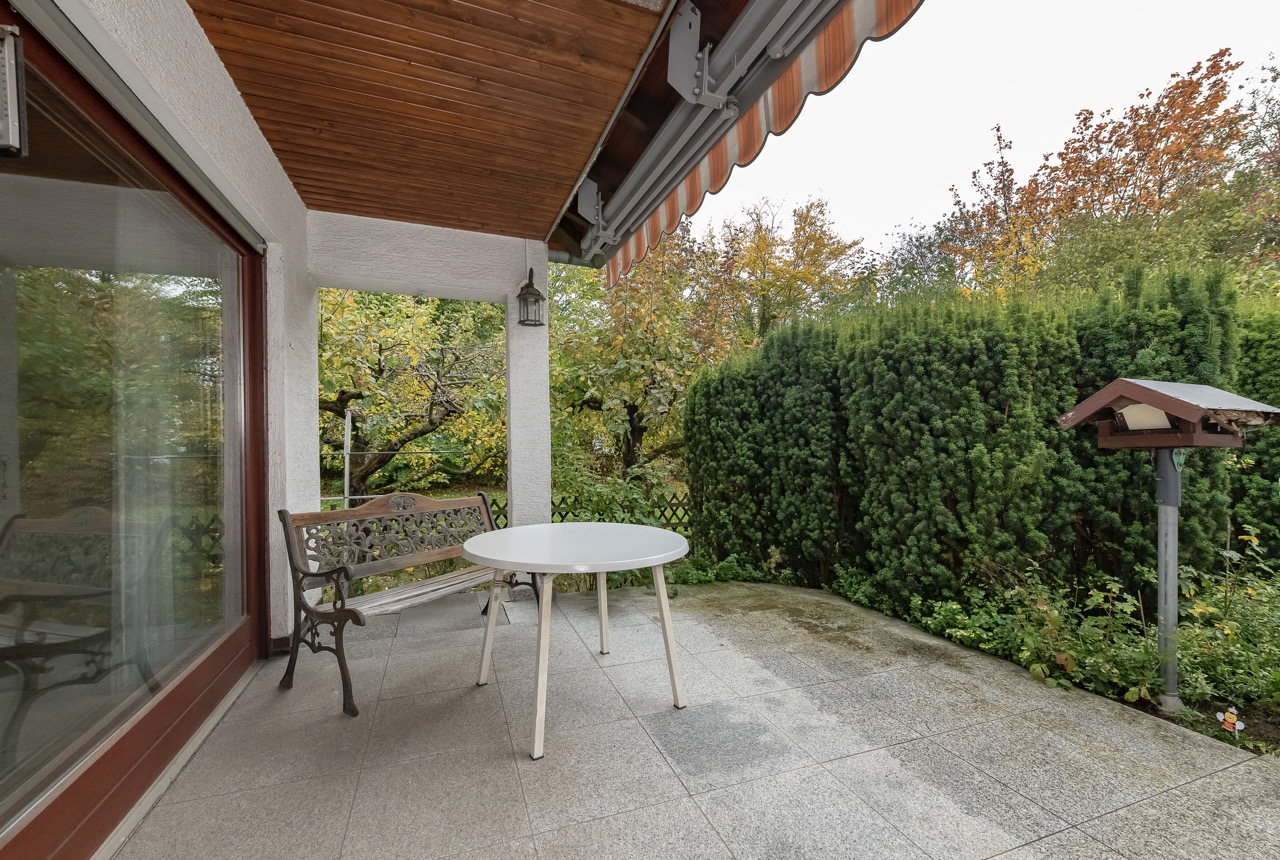 Überdachte Terrasse mit Rundtisch, Bank und Blick auf grüne Hecke und Garten.