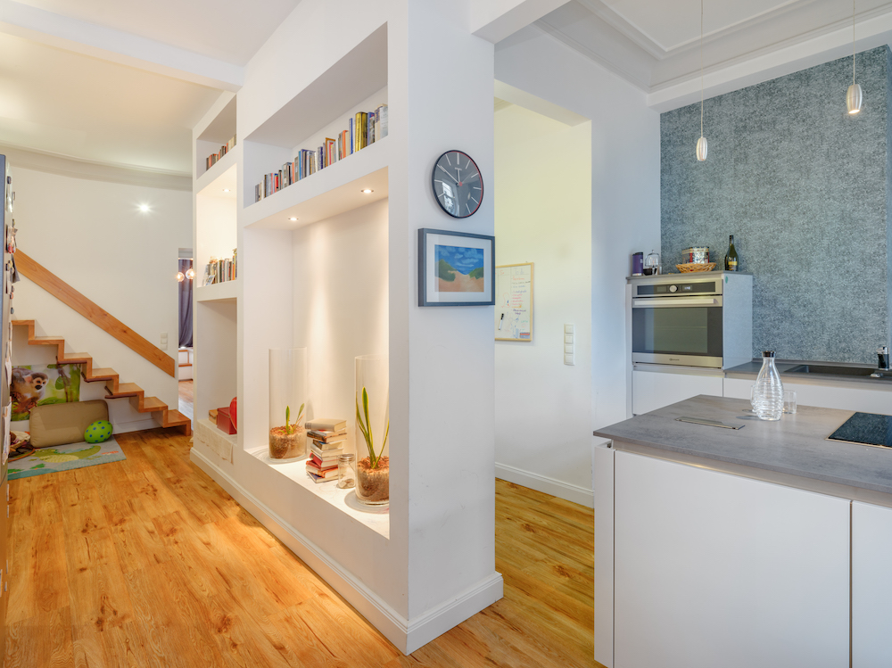 Heller Flur mit Holztreppe, Regal mit Büchern und Pflanzen, angrenzende moderne Küche.