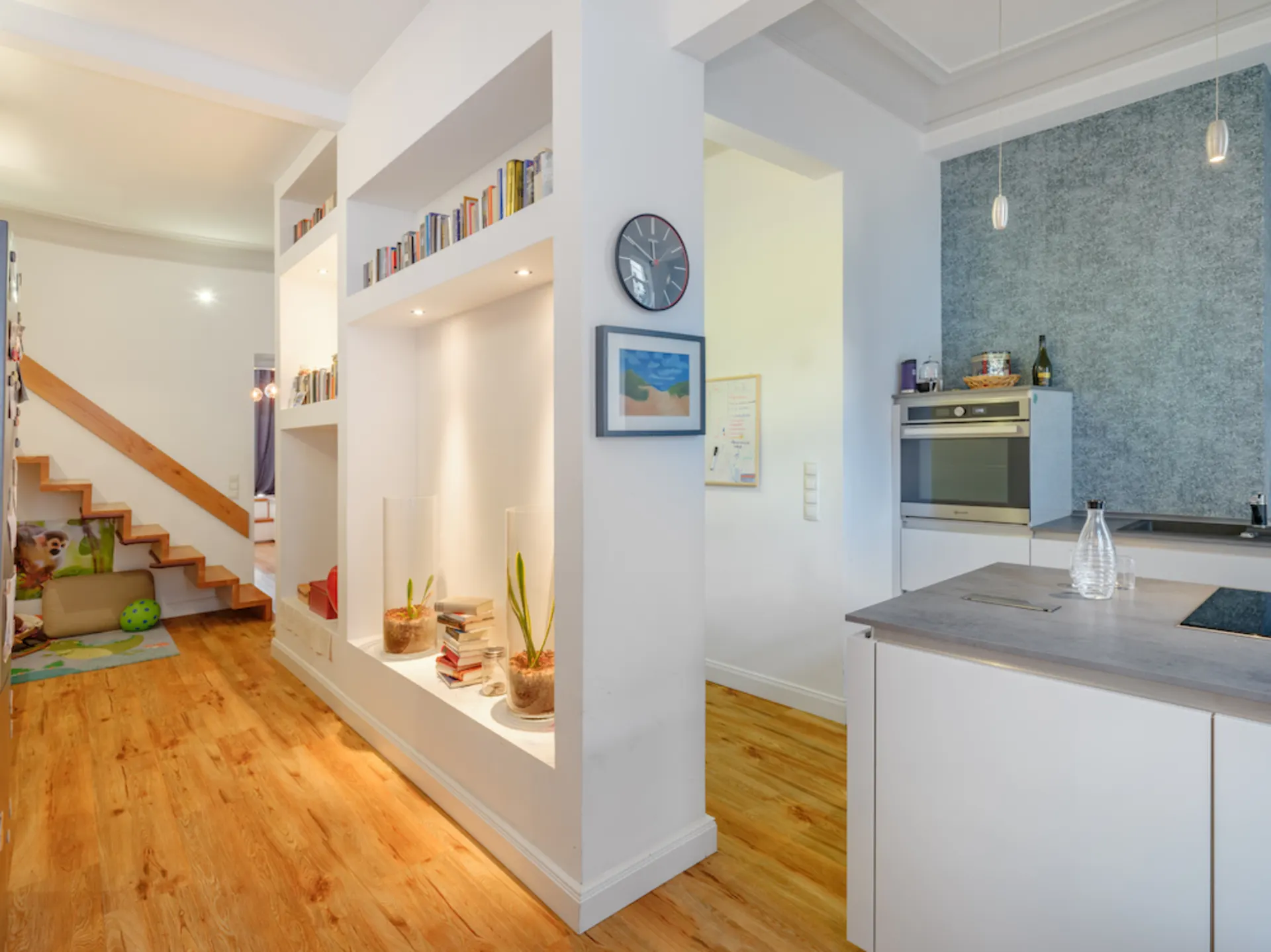 Heller Flur mit Holztreppe, Regal mit Büchern und Pflanzen, angrenzende moderne Küche.