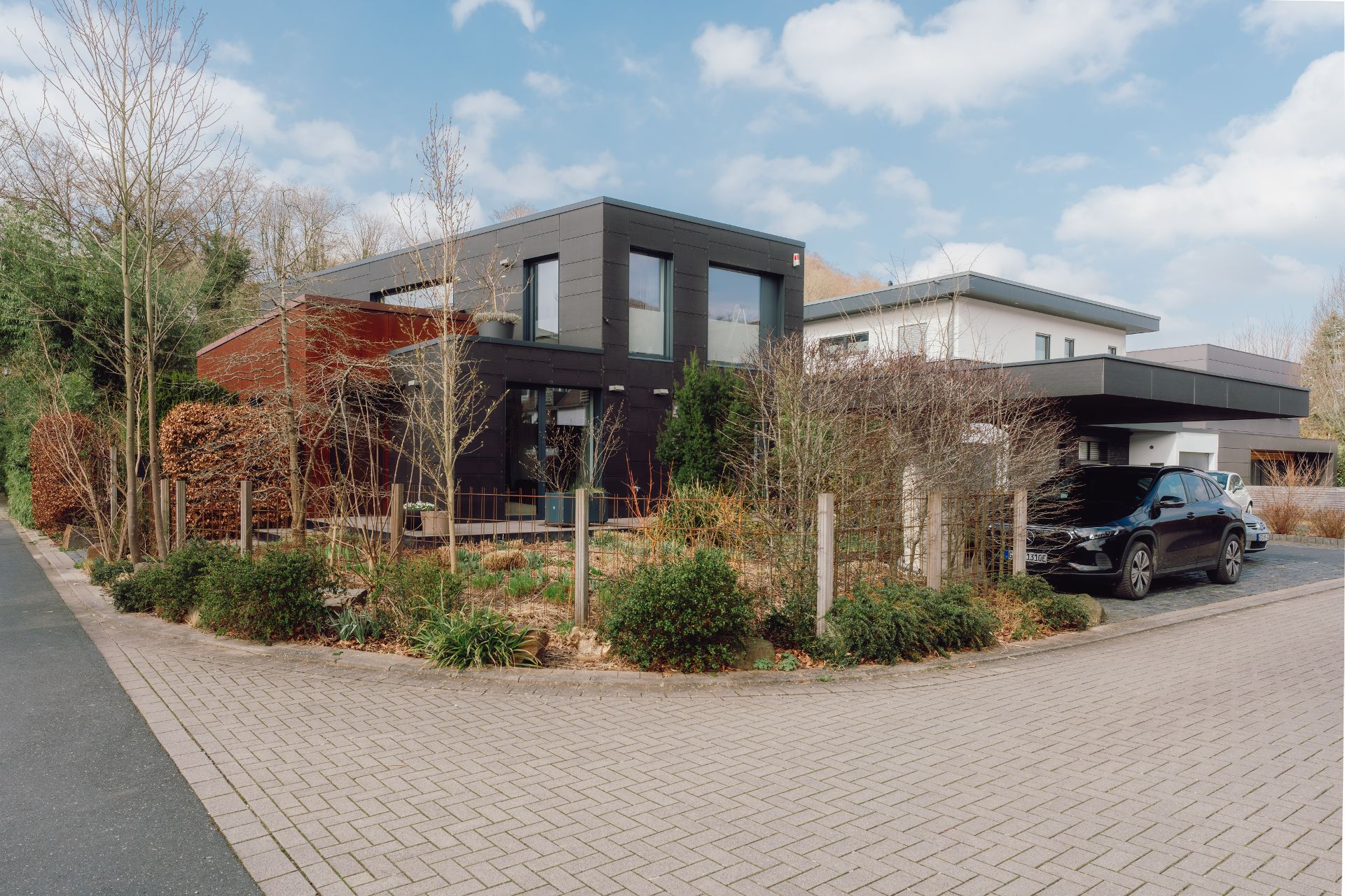 Modernes Wohnhaus mit Flachdach, großem Fenster und Auto vor der Garage, umgeben von Garten und Gebüsch.