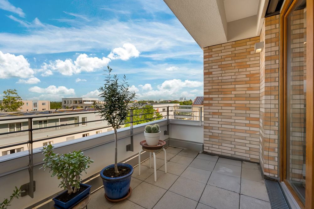 Balkon mit hellen Fliesen, drei Pflanzentöpfen und Blick auf moderne Wohngebäude.