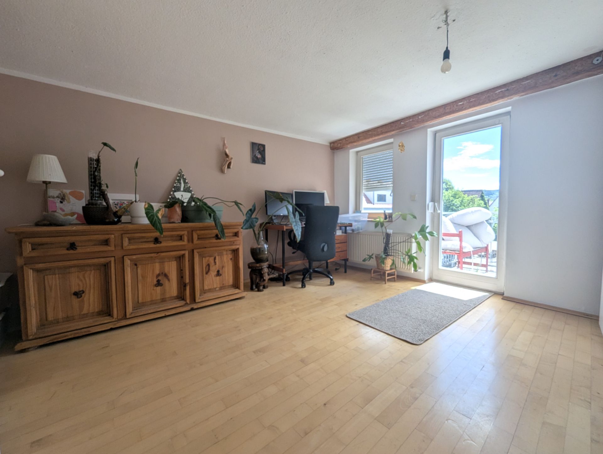 Helles Wohnzimmer mit Holzschrank, Schreibtisch, Bürostuhl und Pflanzen, Tür zum Balkon.