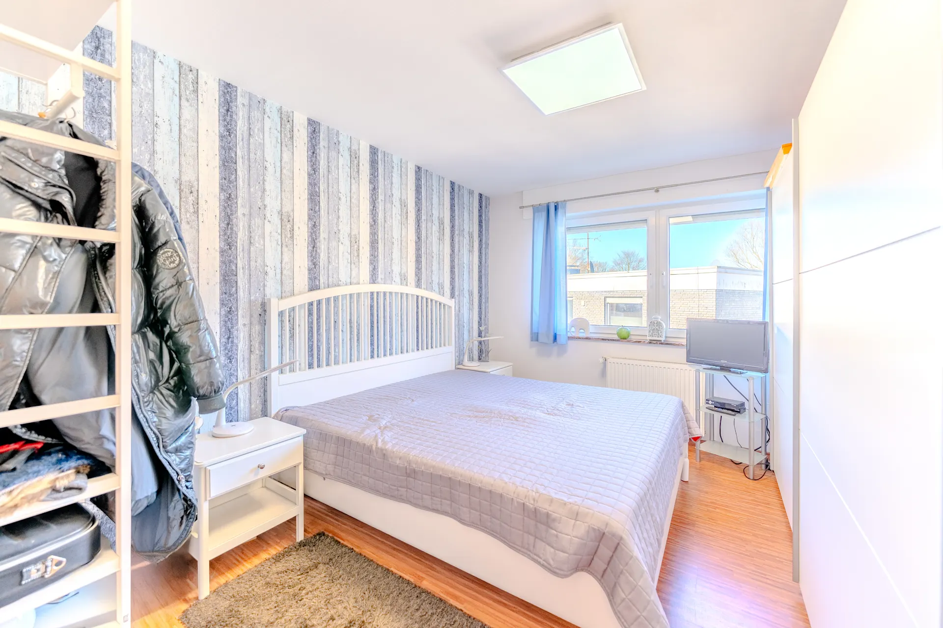 Helles Schlafzimmer mit Bett, Kleiderschrank, Fenster und Fernseher auf kleinem Tisch.