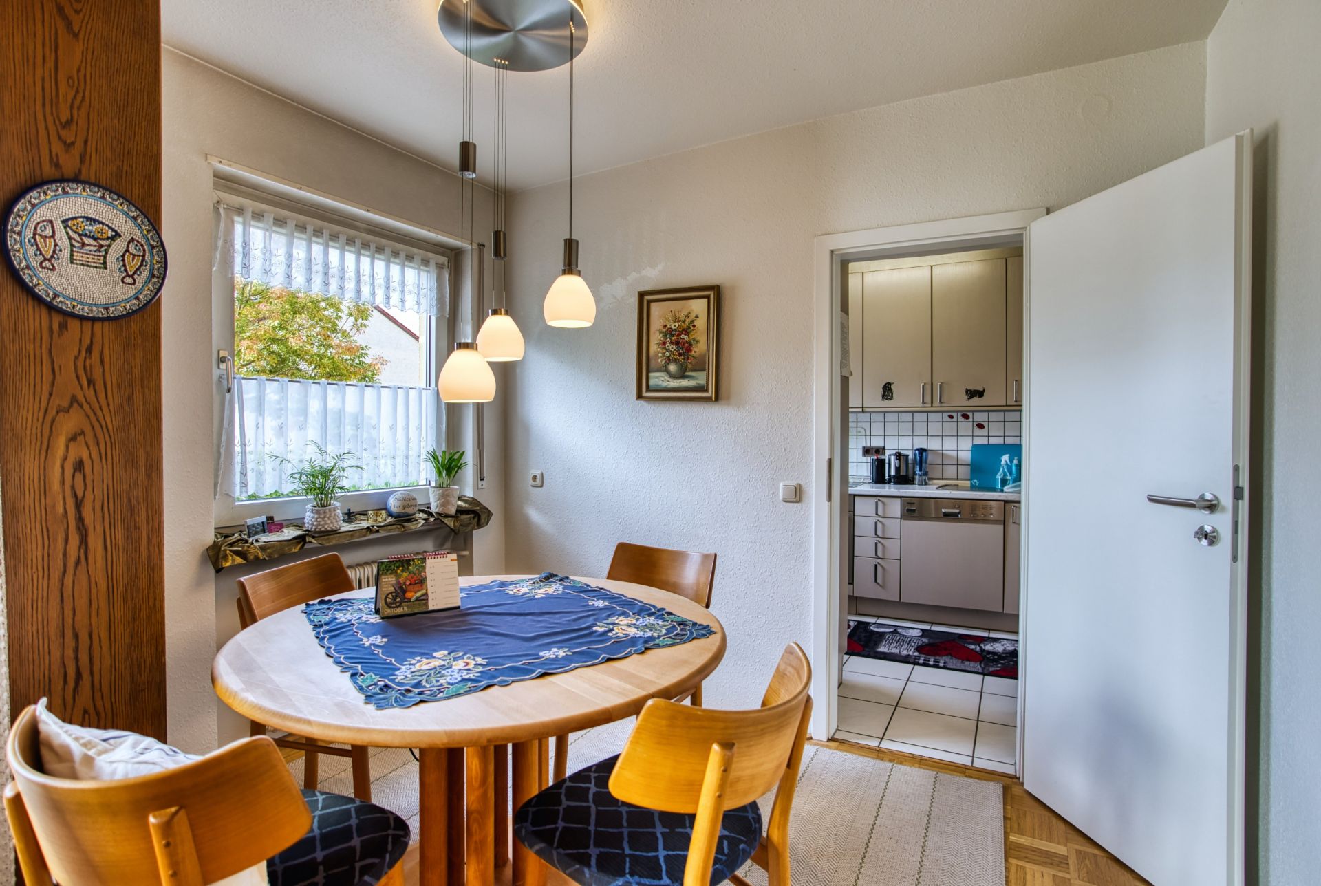 Essbereich mit rundem Tisch, vier Stühlen, Fenster, Wandbild und Blick in die Küche.